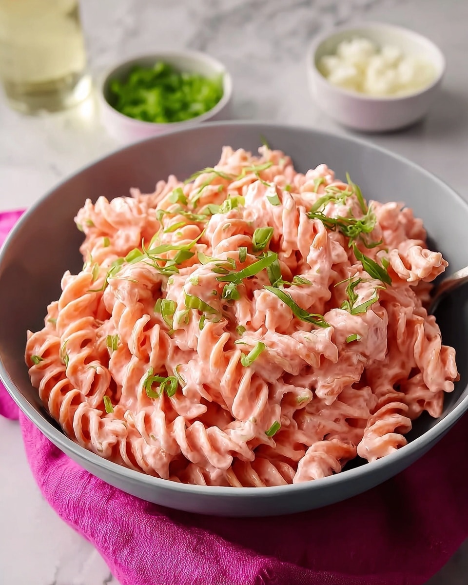 A grey bowl filled with three layers of twisted pasta coated in a thick, creamy pink sauce, topped with small pieces of green herbs scattered on top. The bowl is placed on a bright pink cloth, and two small white bowls with white and green ingredients sit in the blurred background on a white marbled surface. photo taken with an iphone --ar 4:5 --v 7
