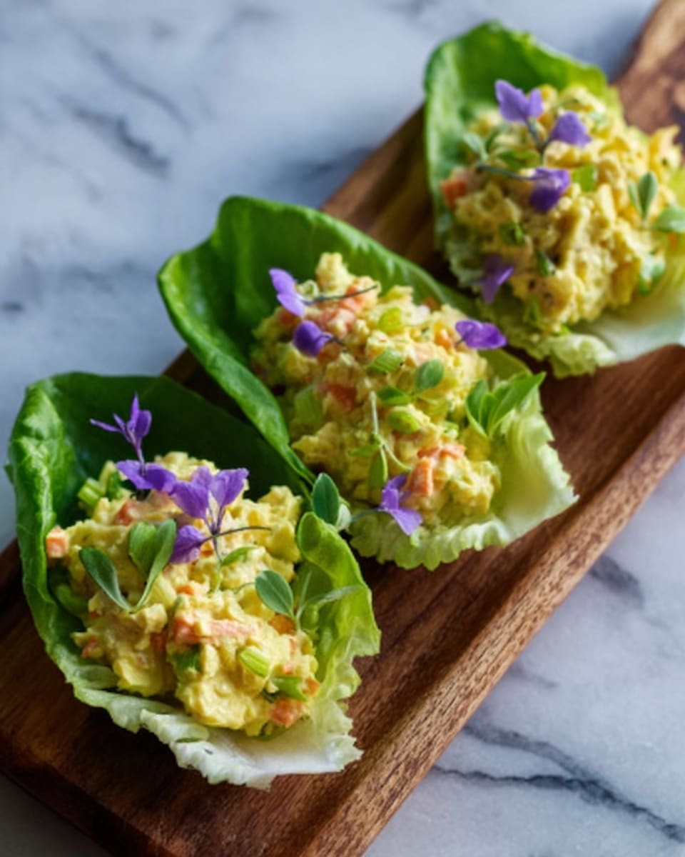 Three small white lettuce cups are placed in a row on a wooden board, each filled with a colorful mixture of chopped ingredients. The filling is mostly light yellow, likely egg salad, with bits of orange carrots and green herbs scattered throughout, topped with thin purple edible flowers and small green leaves. The wooden board rests on a white marbled surface, and the overall look is fresh and tidy. photo taken with an iphone --ar 4:5 --v 7