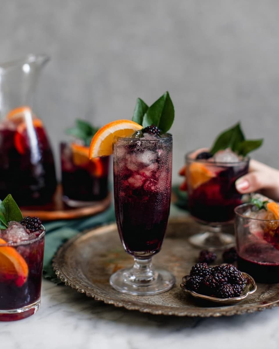 The image shows a tall clear glass filled with a dark purple-red drink. The drink is topped with whole blackberries and pomegranate seeds. On the top edge of the glass, there is a bright orange slice placed at an angle on the right side and a large fresh green leaf resting diagonally across the left side. In the background, two dark-colored coupe glasses with the same dark drink and orange slices can be seen, slightly out of focus. The surface under the glasses is a white marbled texture. Photo taken with an iphone --ar 4:5 --v 7