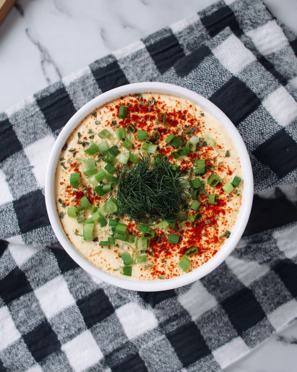 A white bowl filled with a creamy light beige layer topped with scattered bright green chopped scallions, a dusting of red paprika, and finely chopped dark green dill in the center, all placed on a black and white checkered cloth over a white marbled surface. photo taken with an iphone --ar 4:5 --v 7