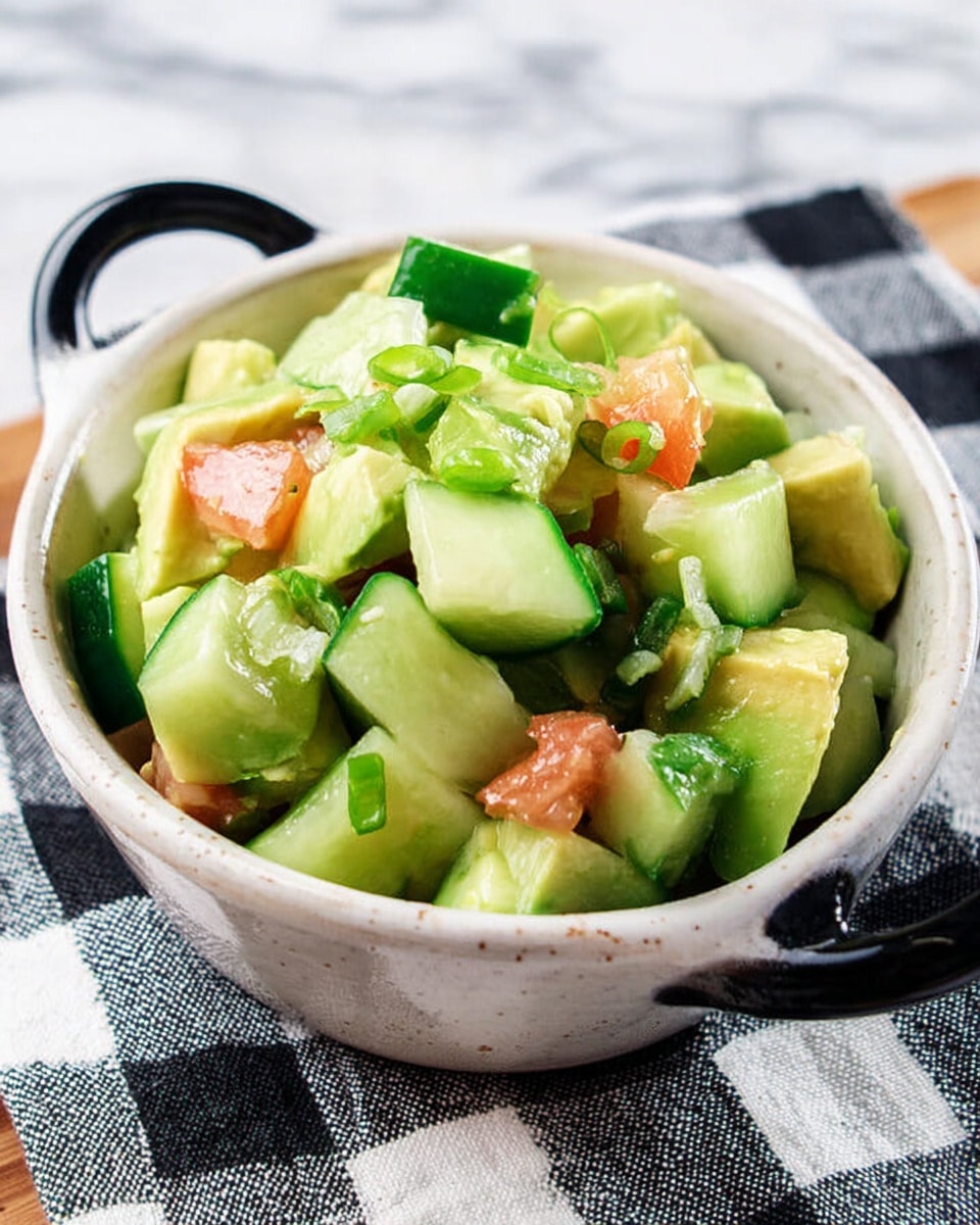 The image shows a small white, speckled bowl with black handles filled with a fresh salad on a black and white checkered cloth over a white marbled background. The salad has three main layers made of chunky pieces of bright green cucumber, light green avocado, and small bits of red tomato, all mixed together. The cucumber pieces are glossy and firm, with some darker green skin visible, while the avocado chunks are creamy with a smooth texture. The tomato pieces add a splash of soft red, with some green scallion slices scattered on top for color and freshness. photo taken with an iphone --ar 4:5 --v 7