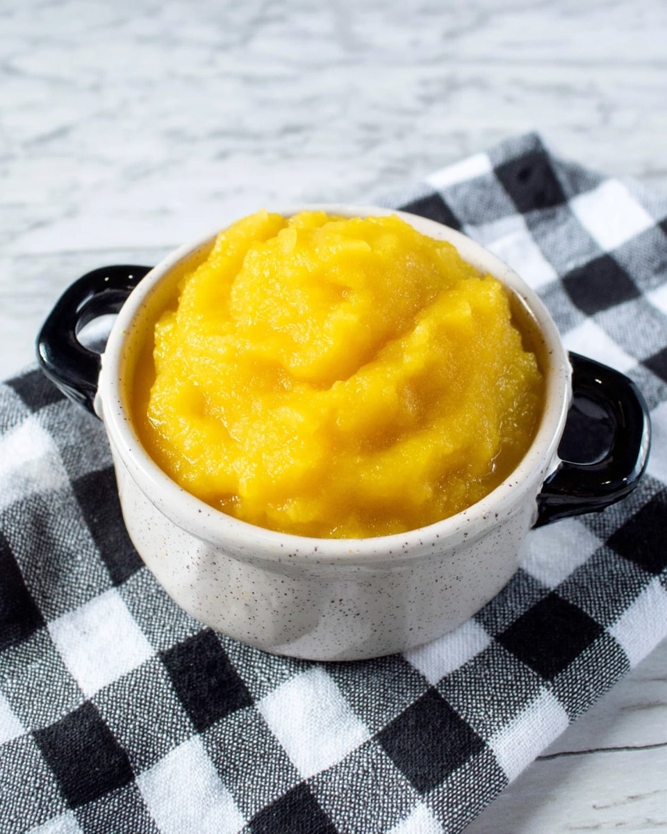 A small white speckled bowl with black handles is filled with a bright yellow, smooth but slightly chunky puree, piled high and slightly mounded above the rim of the bowl. The bowl sits on a black and white checkered cloth, placed on a white marbled surface. The texture of the puree looks soft and moist, with a glossy finish from natural juices. photo taken with an iphone --ar 4:5 --v 7
