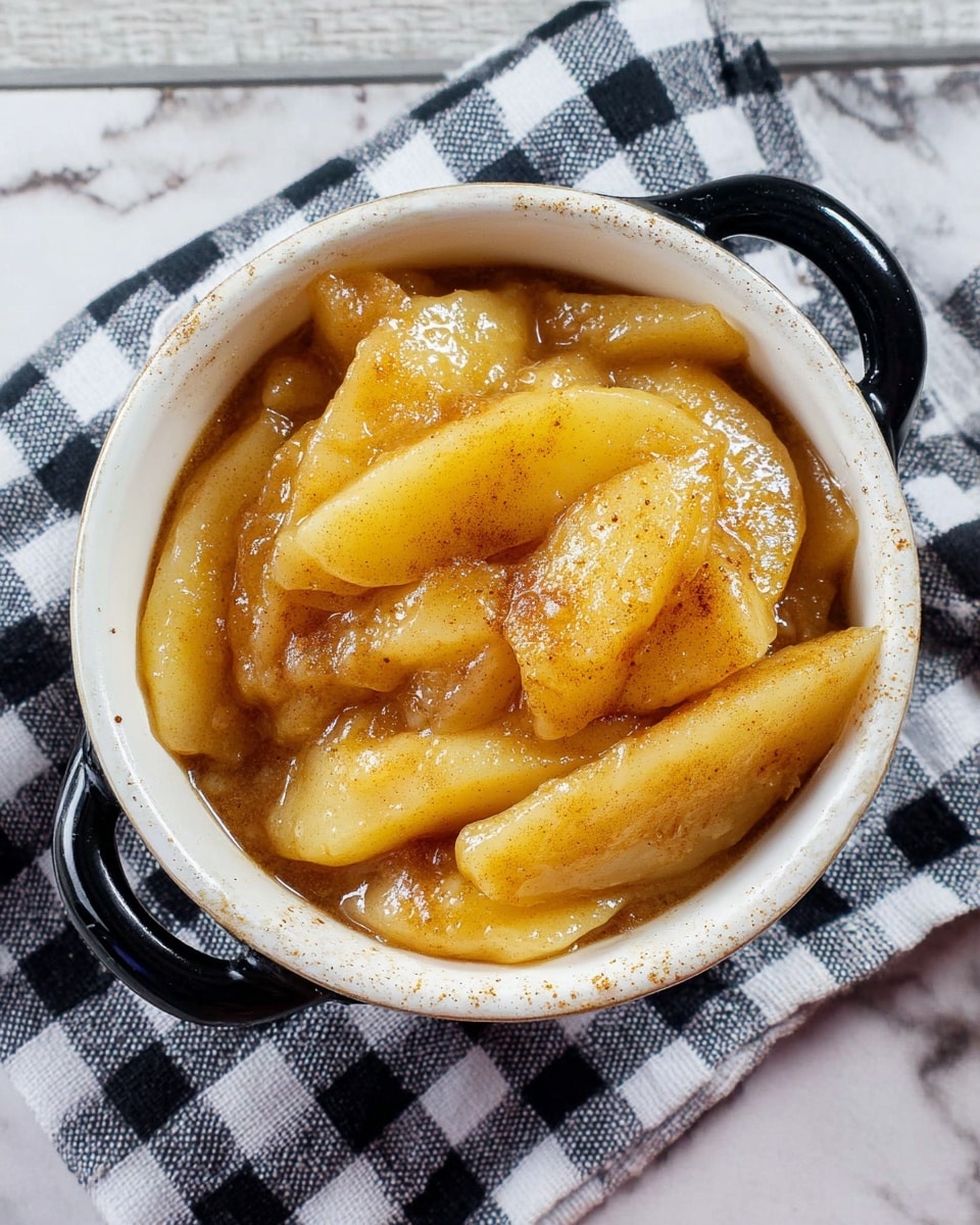 The image shows a small white speckled ceramic pot with black handles filled with cooked apple slices covered in a glossy, thick syrup that is golden brown with visible specks of cinnamon. The apple slices are soft and slightly translucent, stacked unevenly in two to three layers inside the pot. The pot is placed on a black and white checkered cloth over a white marbled surface. The syrup glistens, making the apples look juicy and warm. photo taken with an iphone --ar 4:5 --v 7