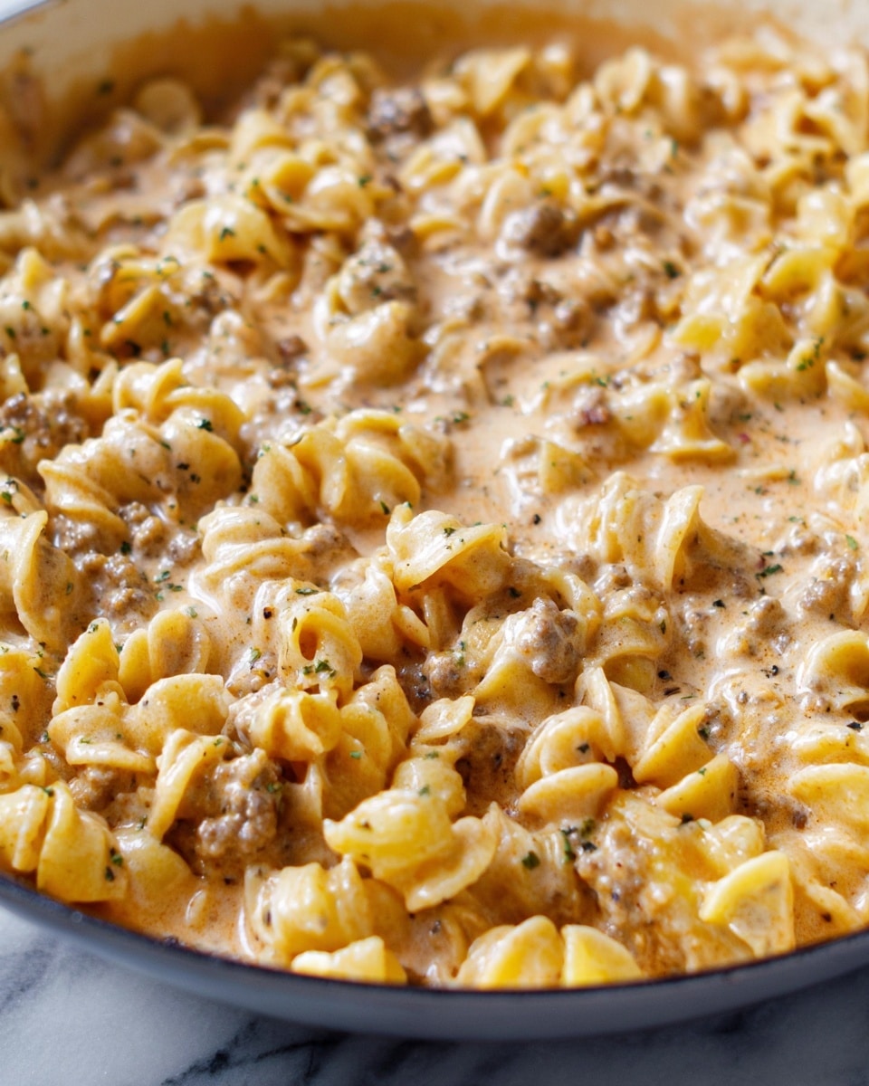 The image shows a close-up view of curvy pasta mixed in a creamy sauce with small bits of ground meat scattered throughout. The pasta is light yellow and soft-looking, coated evenly in a light brown sauce with a smooth, slightly thick texture. Black pepper and small herb flakes are sprinkled on top, adding contrast to the creamy surface. The dish is partially contained in a pan, with the pasta and sauce filling the pan up to its edges, set against a white marbled texture. photo taken with an iphone --ar 4:5 --v 7