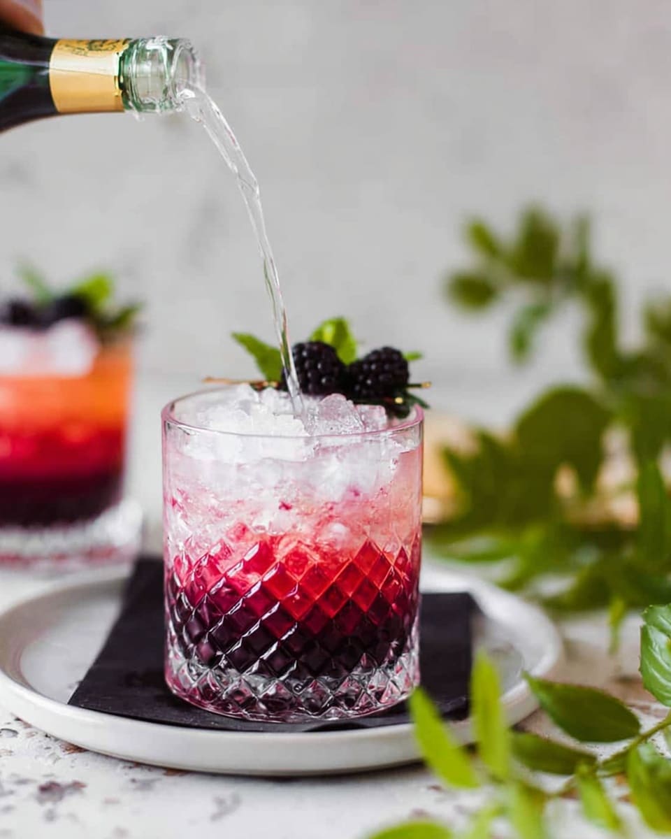 A clear glass with diamond patterns holds a three-layered drink placed on a white plate with a black napkin underneath, all set on a white marbled surface. The bottom layer is deep purple, the middle layer is bright red with a smooth texture, and the top layer is light pink with ice cubes. The drink is garnished on the rim with a small sprig of green leaves and a dark blackberry. A woman’s hand is pouring a clear liquid into the glass from a bottle above. In the background, another identical glass with the same drink and garnish can be seen blurred. There are green leafy branches near the base of the front glass. Photo taken with an iphone --ar 4:5 --v 7