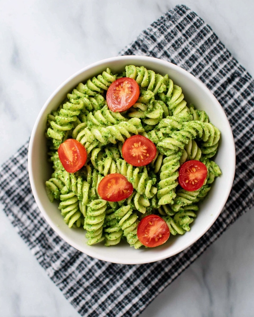A white bowl sits on a black and white checkered cloth over a white marbled surface, filled with green pesto-coated fusilli pasta forming the base layer with its spiral shapes and smooth texture. On top, there are five halved cherry tomatoes scattered evenly, their bright red color and juicy seeds adding a fresh contrast to the green pasta below. The green sauce has a slightly coarse texture, coating each pasta piece well, while the overall presentation looks fresh and vibrant. Photo taken with an iphone --ar 4:5 --v 7