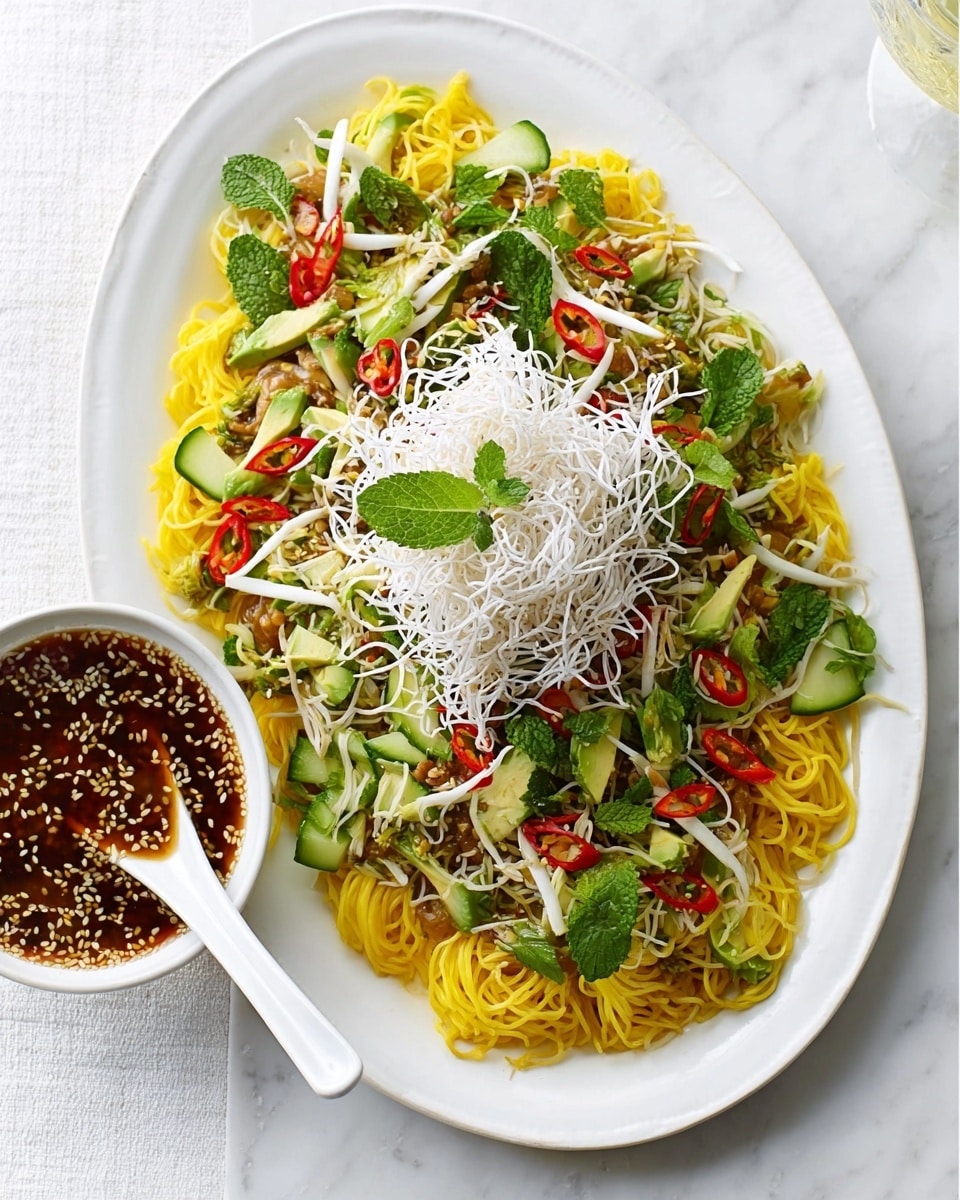 The dish is presented on a white oval plate set on a white marbled textured surface. The bottom layer is made of yellow noodles spread across the plate, topped with a mix of fresh green herbs and light green avocado slices that add a soft texture. Bright yellow and red thinly sliced vegetables, possibly bell peppers, are scattered evenly among the greens. On top sits a generous layer of crispy white fried noodles, giving a crunchy contrast. A white bowl nearby contains a dark sauce with visible sesame seeds, and a white spoon rests in it. The scene has a fresh and colorful look. Photo taken with an iphone --ar 4:5 --v 7