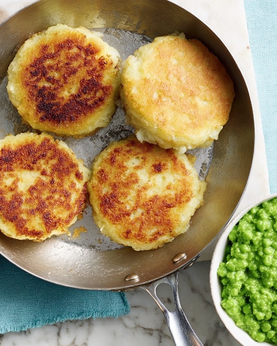 A silver pan contains four round, golden-brown cakes with a crispy outside and soft inside texture. The cakes are arranged with some space between them, showing a slightly uneven, crumbly surface with browned spots from cooking. Below the pan is a white marbled surface. In the lower left corner, there is a white bowl filled with bright green, mashed peas. The overall scene suggests a simple and warm cooking moment. Photo taken with an iphone --ar 4:5 --v 7