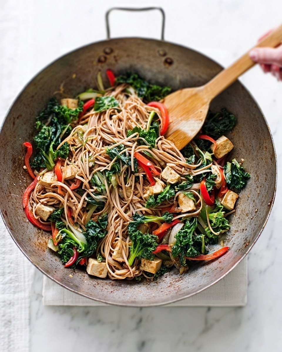 A rustic wok filled with a colorful stir-fry sits on a white marbled surface. The dish has three main layers: a base of light brown soba noodles with a smooth texture, topped with bright green kale leaves that look fresh and slightly wilted, and scattered pieces of light golden brown tofu with a firm texture. Thin strips of red bell pepper add a splash of vibrant red across the dish. A wooden spatula and a woman's hand holding a wooden spoon dip into the noodles, adding a sense of action. The wok has a weathered metal look with a wooden handle. Photo taken with an iphone --ar 4:5 --v 7