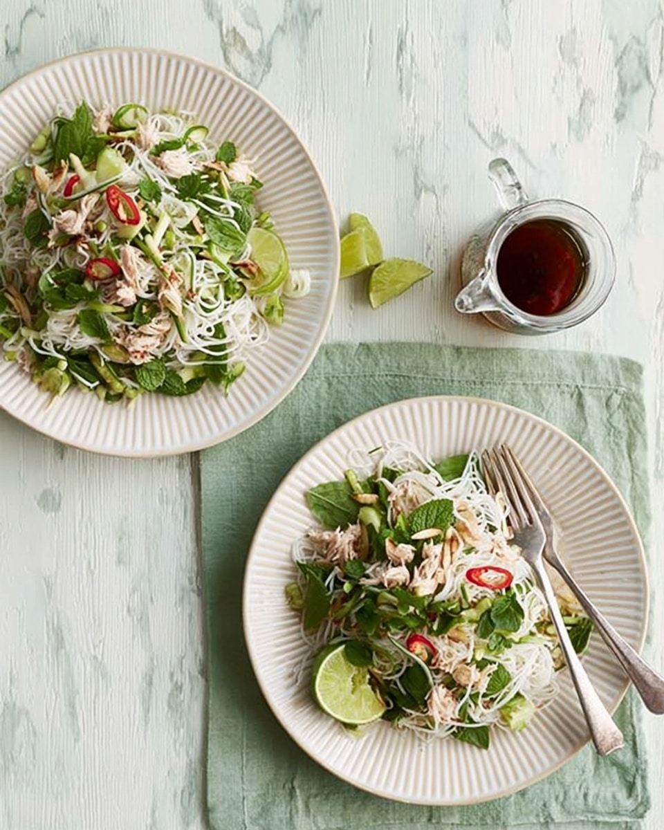 Two white plates are placed on a light green cloth over a white marbled surface, each filled with a fresh salad. The salad has a base of green leafy vegetables mixed with white shredded ingredients, possibly shredded chicken or cheese, and thin white noodles or bean sprouts on top. There are small red bits scattered throughout the salad adding a touch of color. Each plate has a wedge of lime placed at the edge. A clear glass bottle with dark liquid sits near the top left plate. Two silver forks lie next to the bottom plate. photo taken with an iphone --ar 4:5 --v 7