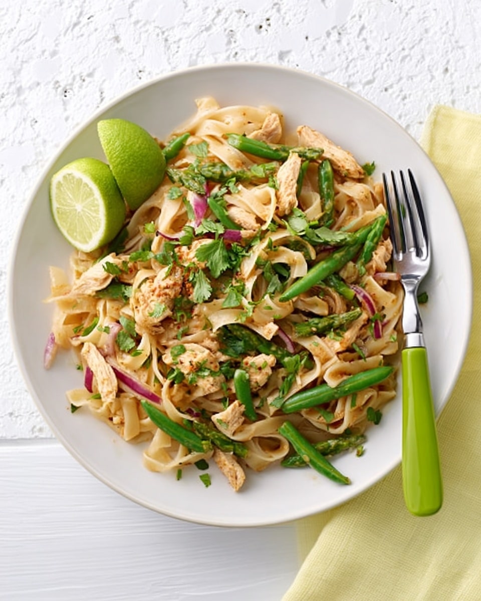 A white bowl filled with a noodle dish that has three main layers: wide, flat beige noodles forming the base, thin light brown strips of cooked chicken on top, and scattered bright green herbs and chopped green beans layered throughout for freshness. There are small bits of red onion adding a touch of purple color mixed in. A halved lime with a pale green inside is placed at the upper left edge of the bowl. The bowl sits on a folded pale yellow napkin on a white marbled surface. To the right of the bowl, there is a silver fork with a pale green handle. Photo taken with an iphone --ar 4:5 --v 7