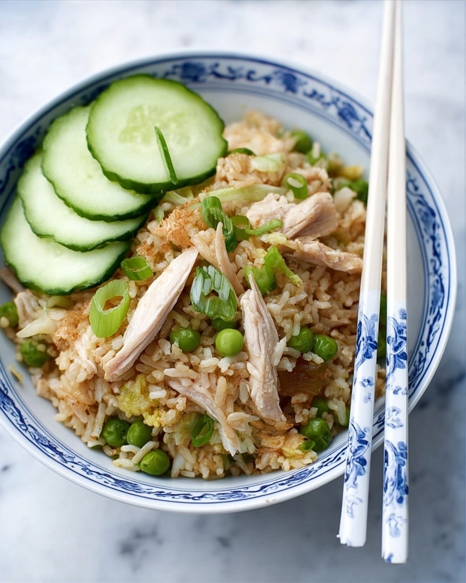 The image shows a white bowl with blue patterns filled with fried rice. The fried rice has a light brown color with visible pieces of green peas and green onion slices mixed throughout. On top of the rice, there are several slices of light brown cooked chicken and three slices of green cucumber placed on one side. Two white chopsticks with blue patterns rest on the side of the bowl. The bowl is sitting on a white marbled surface. Photo taken with an iphone --ar 4:5 --v 7