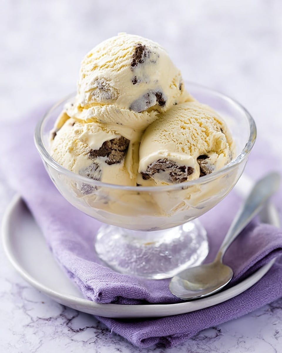 A clear glass bowl sitting on a white saucer with a silver spoon rests on a folded lavender cloth over a white marbled surface. Inside the bowl, there are three scoops of creamy vanilla ice cream with visible chunks of dark chocolate mixed in. The ice cream has a smooth yet slightly melting texture, and the chocolate pieces are irregularly shaped, scattered throughout each scoop. The light purple backdrop adds a soft contrast to the pale tones of the ice cream and the clear glassware, making the dessert look inviting and fresh. photo taken with an iphone --ar 4:5 --v 7