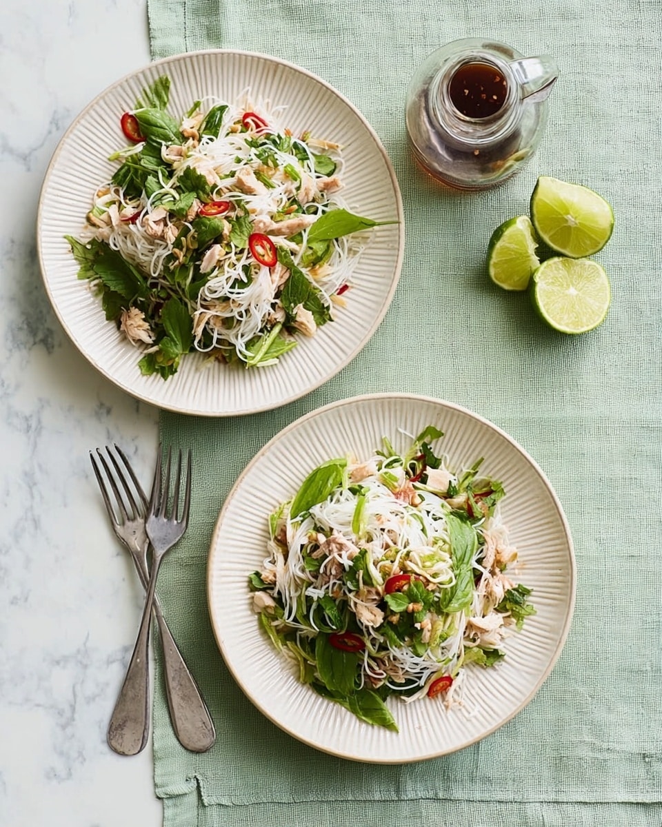 Two white round plates with ridged edges, each filled with a layered salad. The base layer is light green leafy greens, topped with white thin noodles, small pieces of light brown shredded chicken, red chili flakes scattered across, and some green herbs. Each plate has a small lime wedge on the side. One plate has a silver fork and spoon placed next to it on a light green cloth. Above the cloth, there is a small glass bottle with a brown liquid. All items are on a white marbled surface. Photo taken with an iphone --ar 4:5 --v 7