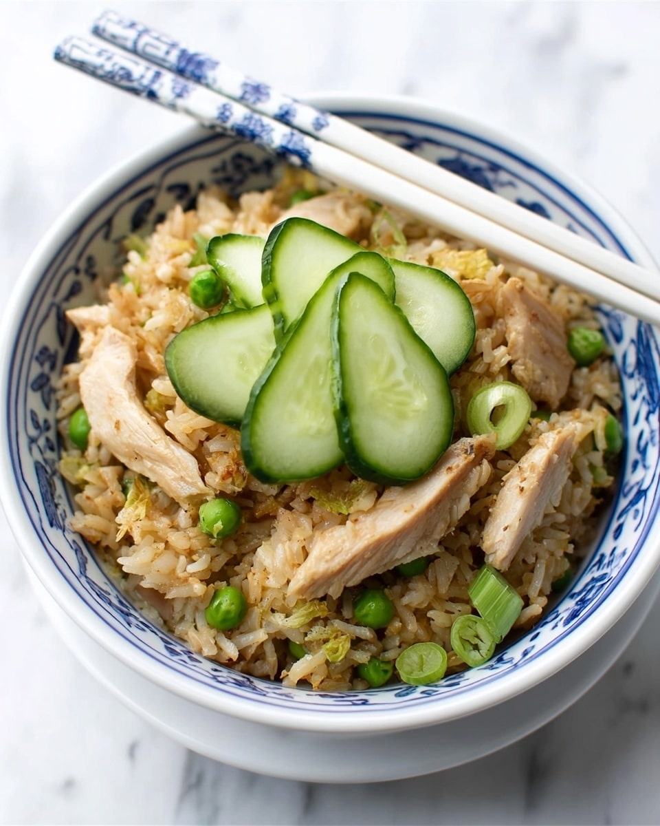 A white bowl with blue patterns contains a layered fried rice dish. The bottom layer is light brown fried rice mixed with small green peas and pieces of chopped green onion. On top of the rice are large slices of light brown cooked chicken and three crescent-shaped slices of fresh green cucumber placed at the edge. A pair of white chopsticks with blue designs rests on the side of the bowl. The bowl is placed on a white marbled surface. Photo taken with an iphone --ar 4:5 --v 7