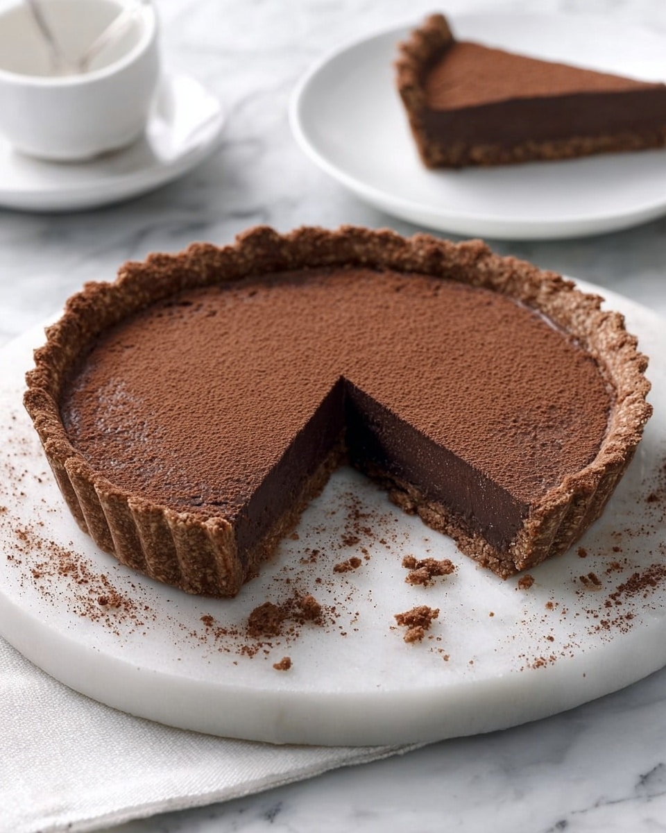 A round tart sits on a white marble tray, with one slice removed to show two distinct layers inside: a thick, crumbly, light brown crust on the outside and a smooth, dark chocolate filling dusted with fine cocoa powder on top. The tart crust is rough textured and holds the soft filling firmly. In the background, blurred on a white plate, the removed slice rests next to a white bowl on a white marbled surface, creating a clean and bright setting. Photo taken with an iphone --ar 4:5 --v 7