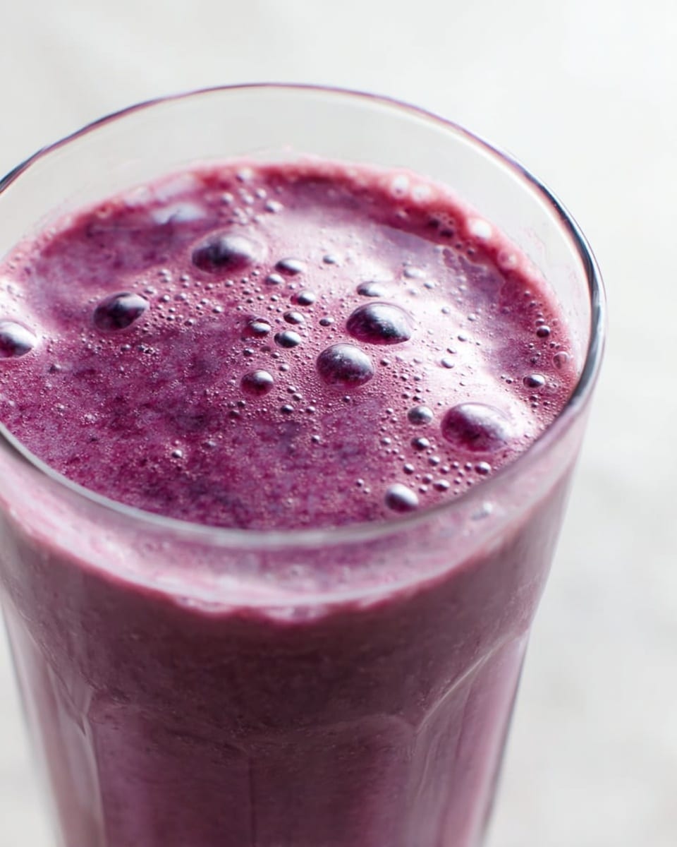 A close-up of a purple smoothie inside a clear glass, showing a thick, slightly foamy texture with many small bubbles on the surface, giving it a fresh and blended look. The background is a white marbled texture. Photo taken with an iphone --ar 4:5 --v 7