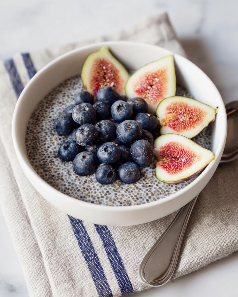 A white bowl filled with a creamy chia pudding, speckled with tiny black chia seeds throughout the light grayish base. On top, there are four slices of fig with pale green outer skin and deep pinkish-red inside with small seeds, placed evenly around the center. In the center, there is a cluster of fresh blueberries with a deep blue color, creating a contrast with the fig slices and chia pudding. The bowl sits on a beige textured cloth with a gray stripe, on a white marbled surface, and there is a shiny silver spoon near the top left side. photo taken with an iphone --ar 4:5 --v 7