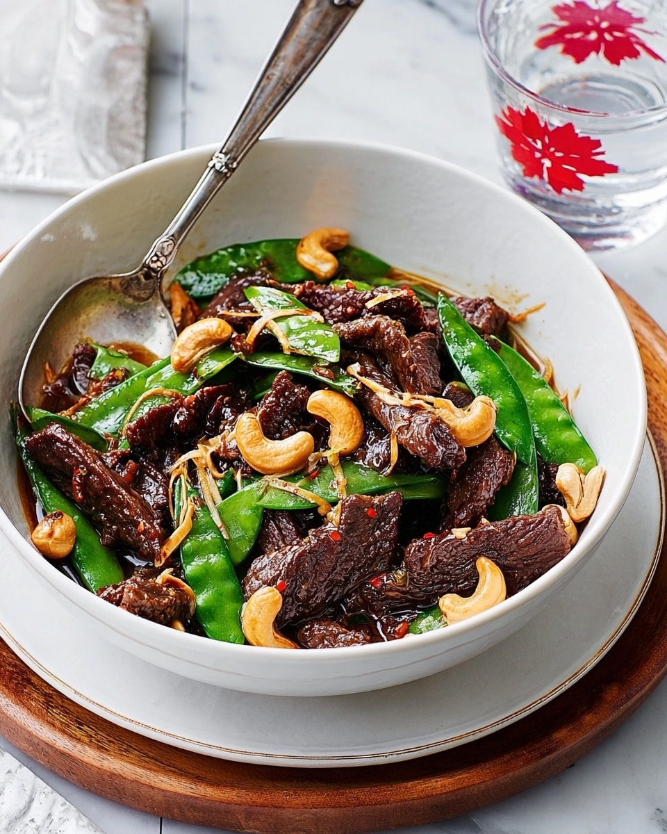 A white bowl holds a stir-fry with about three layers: the bottom layer has dark brown sauce coating tender strips of beef, above that are bright green snow peas and golden brown cashew nuts scattered around, and thin light brown slivers of ginger lay on top. A white spoon stands inside the bowl, partially covered with the sauce and vegetables. The bowl sits on a white marbled surface with a round white plate beneath it edged with thin gold trim. Nearby, a glass of clear water with red flower designs is partially visible. Photo taken with an iphone --ar 4:5 --v 7