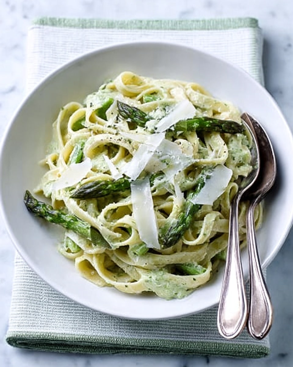 A white plate holds a creamy pasta dish, with wide fettuccine noodles coated in a smooth white sauce. Pieces of green asparagus are mixed in and scattered on top. Thin, irregular shavings of pale yellow cheese rest on the pasta, while a sprinkle of black pepper adds small dark specks. A fork and knife made of shiny metal are placed crossed on the right side of the plate. The plate sits on a folded white cloth with light blue and green stripes, all set on a white marbled surface. photo taken with an iphone --ar 4:5 --v 7