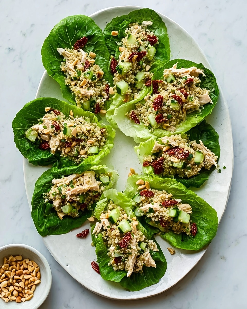 A white oval plate on a white marbled surface holds six green leafy lettuce cups as the base layer. Each cup is filled with a mix of light green couscous or quinoa grains, small chunks of white chicken, and diced pale green avocado. There are sun-dried tomato pieces adding a reddish-brown color on top. Small light tan pine nuts are scattered throughout the filling. Some fresh green herbs are placed on top of each cup, adding a touch of bright green detail. A small white bowl of pine nuts is visible near the plate. Photo taken with an iphone --ar 4:5 --v 7