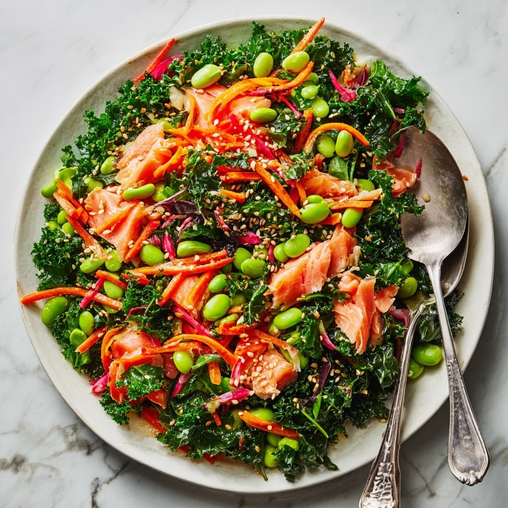A large white plate filled with a colorful salad featuring three main layers: the base layer is bright green curly kale leaves spread all over, the middle layer consists of small green edamame beans scattered evenly, and the top layer has pieces of pink cooked salmon mixed with thin orange carrot strips and red bell pepper slices. The salad is sprinkled with light brown sesame seeds, and there are two metal utensils—one spoon and one fork—resting on the right side of the plate with some salad on them. The background is a white marbled texture. photo taken with an iphone --ar 4:5 --v 7