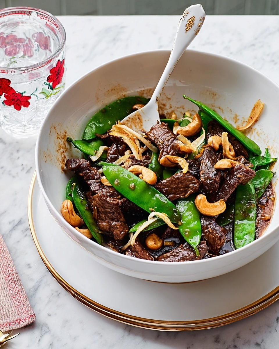 A white bowl filled with several layers of a cooked dish sits on a white marbled surface. The bottom layer shows dark brown strips of beef, cooked with a slightly glossy texture. Spread among the beef are bright green snap peas giving a fresh pop of color. Scattered over and around the beef and peas are golden cashew nuts, adding a bit of crunch. Thin light brown slices that look like chopped garlic or ginger are mixed in, showing fine texture and detail. A silver spoon rests in the bowl, leaning against the side. The white bowl sits on a wooden tray with a round white plate underneath. A glass of water with a red flower design sits in the background on the right side. Photo taken with an iphone --ar 4:5 --v 7