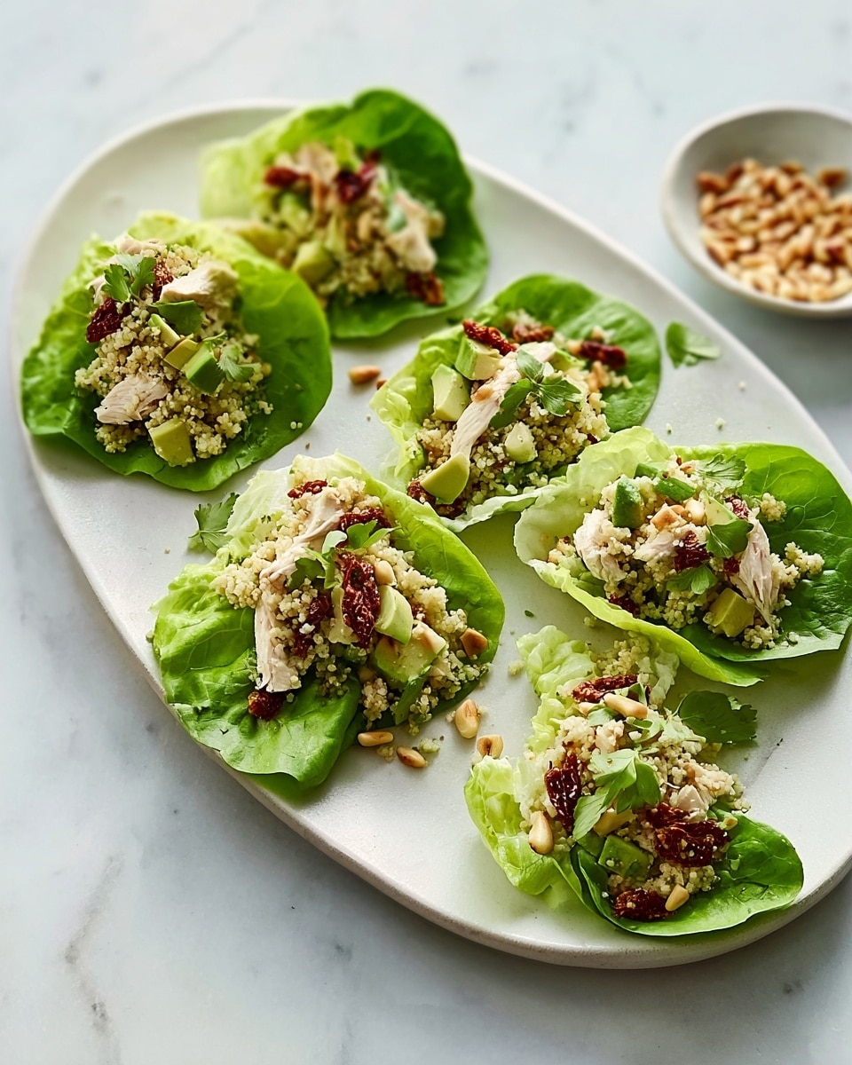 An oval white plate on a white marbled surface holds six large, deep green lettuce leaves arranged neatly. Each leaf is topped with a mix of small beige quinoa grains, green chopped cucumber pieces, shredded light brown chicken, small bright red sun-dried tomatoes, and sprinkled pine nuts. The mixture looks fresh and colorful against the dark green lettuce. A small white bowl with extra pine nuts sits near the plate. The overall look is light and healthy, with a fresh, natural feel. photo taken with an iphone --ar 4:5 --v 7