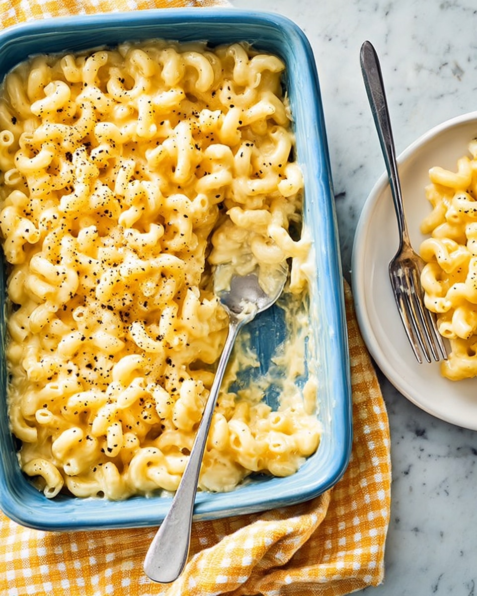 A close-up shot of a baked macaroni and cheese dish in a white rectangular casserole dish on top of a yellow checkered cloth. The macaroni is creamy light yellow, coated with smooth melted cheese, and sprinkled with black pepper. A serving spoon is partly inside the dish, scooping macaroni. To the right, a white round plate holds a small portion of the macaroni and cheese with a silver fork resting beside it. The background is a white marbled texture. Photo taken with an iphone --ar 4:5 --v 7