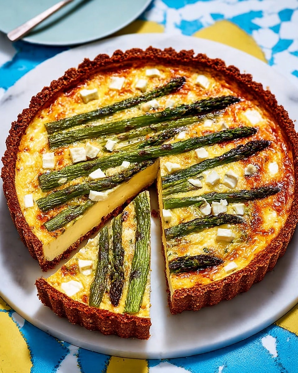 The image shows a round asparagus tart with a golden-brown crust that is slightly thick and crispy on the outer edge. Inside the tart, the bottom layer is a creamy, light yellow cheese filling with small white cheese cubes scattered throughout. On top, there are whole green asparagus spears arranged evenly across the tart, their texture slightly glossy and roasted with some browned spots. One wedge-shaped slice is cut and partly lifted, showing the layers clearly. The tart is placed on a white plate with a smooth matte finish, set on a white marbled surface with a blue and yellow pattern partially visible. Photo taken with an iphone --ar 4:5 --v 7