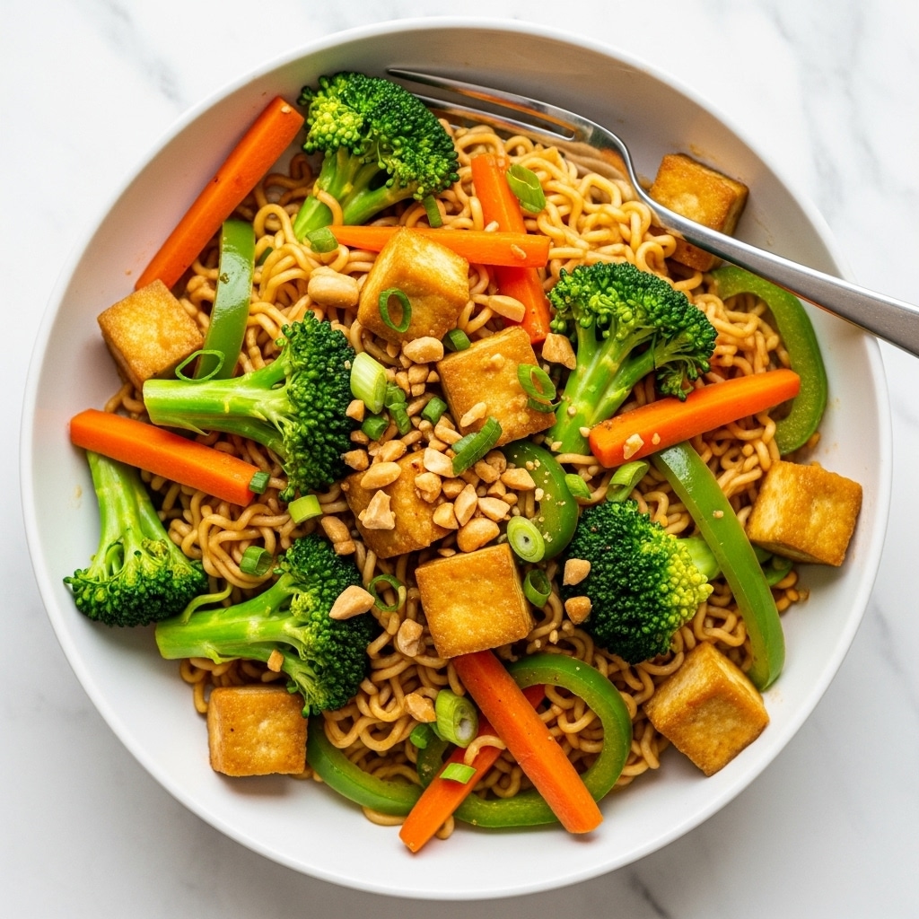 A white plate holds a colorful stir-fry of instant noodles mixed with various vegetables and tofu. The bottom layer is twisted, light brown noodles, covered in small chopped green onions scattered throughout. On top of the noodles, there are bright green broccoli florets, thin orange carrot sticks, and slices of red bell pepper, along with thick cubes of golden-brown tofu that show a slightly crisp texture. Some peanuts are sprinkled over the dish, adding a crunchy texture, and a shiny sauce lightly coats everything, giving a slight glaze. A silver fork rests on the edge of the plate, and the whole scene is set on a white marbled surface. Photo taken with an iphone --ar 4:5 --v 7