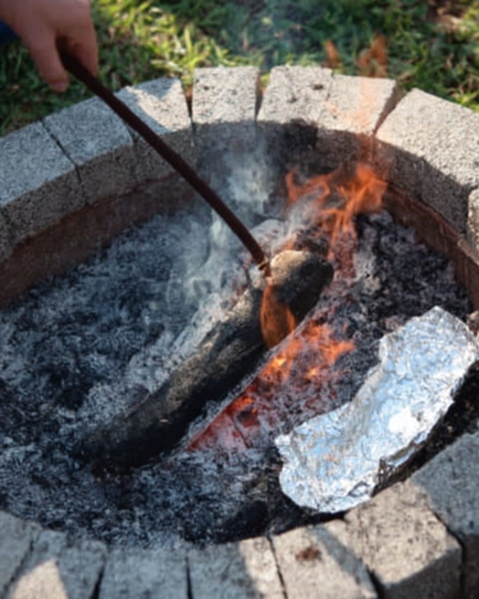The image shows a blackened log burning over glowing red and orange flames, surrounded by thick white and gray ashes. There is a piece of shiny silver foil resting on the edge of the fire pit, which is built with bricks and concrete. A woman's hand is visible holding a thin metal stick near the fire. The background surface has a white marbled texture. Photo taken with an iphone --ar 4:5 --v 7