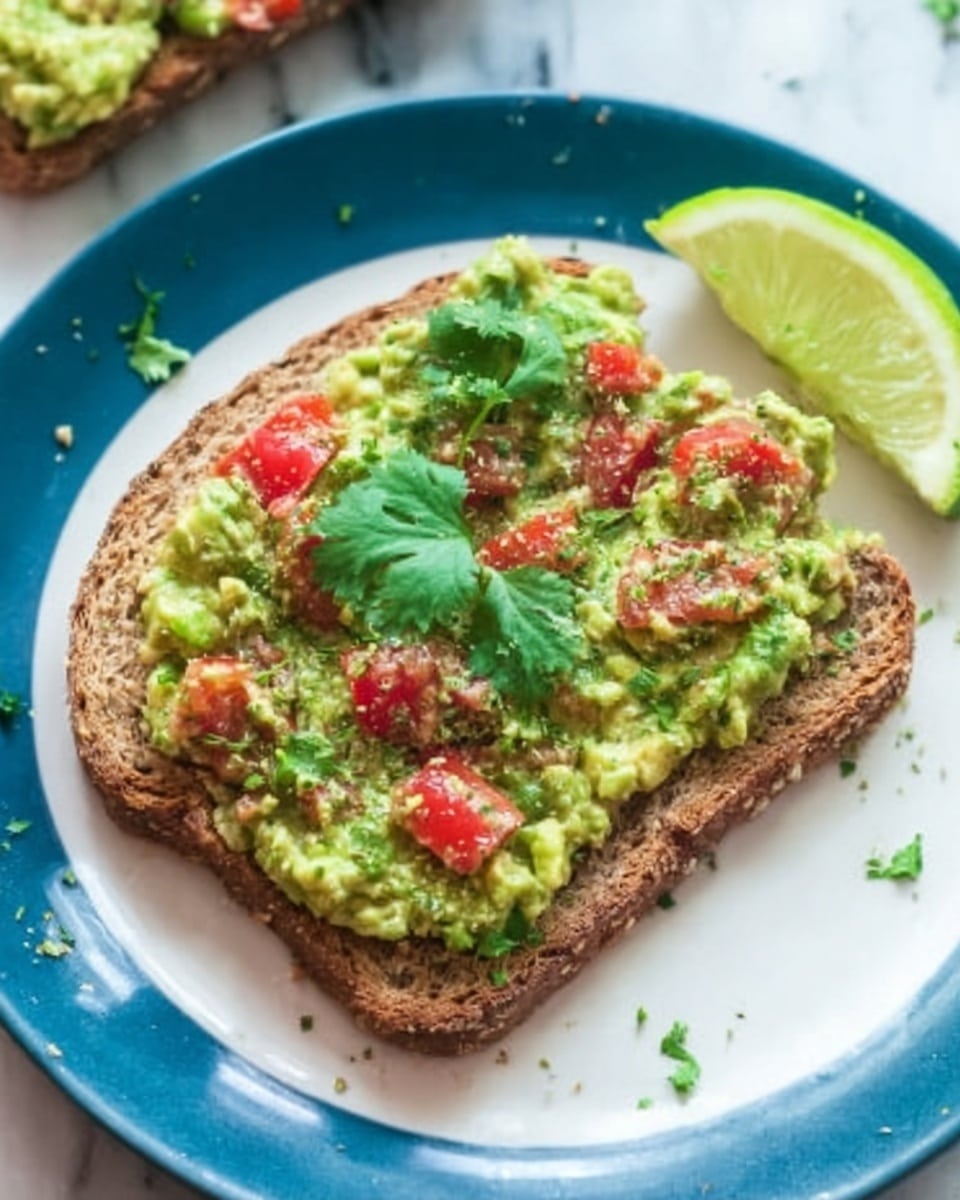 The image shows a blue plate with two slices of multigrain bread topped with chunky mashed avocado mixed with small pieces of tomato and garnished with fresh cilantro leaves. The avocado layer is thick and spread evenly across each bread slice, showing a textured mix of green and red colors. A wedge of lime sits on the side of the plate, and there is a white marbled surface beneath the plate. A woman's hand is holding a spoon above the plate, with some green herbs scattered around. Photo taken with an iphone --ar 4:5 --v 7
