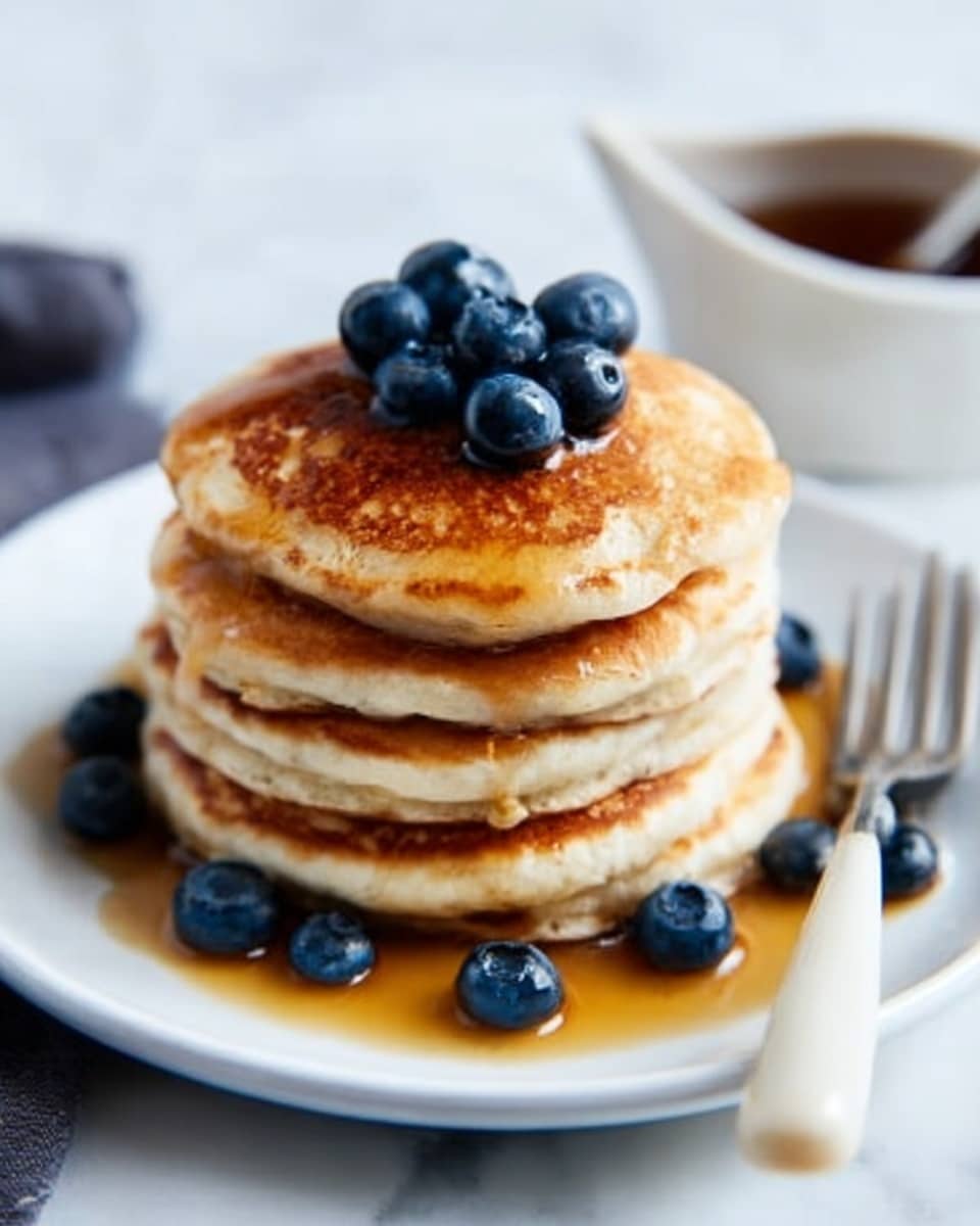 A stack of five golden brown pancakes sits in the center of a white plate on a white marbled surface. The pancakes are thick and fluffy, with a smooth, slightly crispy top layer. On the top pancake, there is a small pile of fresh blueberries, deep blue with a slight shine. Some syrup is poured over the top pancake, dripping down the sides and pooling lightly at the base, shining in the light. Around the plate, a few more blueberries rest near the pancakes. In the background, a small white cup of syrup and a fork are slightly blurred. Photo taken with an iphone --ar 4:5 --v 7