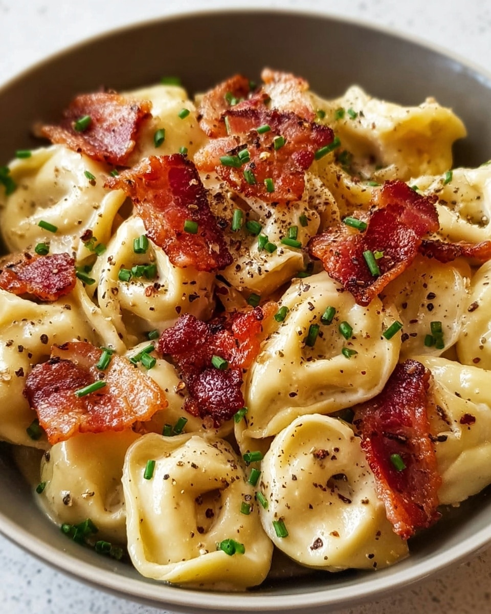 A white bowl filled with two layers of creamy tortellini pasta coated in a light cheese sauce, each tortellini round and smooth with a glossy texture. On top, there is a layer of crispy bacon pieces in a deep reddish-brown color, scattered unevenly over the pasta. Small green chopped chives are sprinkled generously on all layers, along with broken black pepper flakes adding specks of black and texture contrast. The dish looks rich and has a warm color palette against a white marbled background. Photo taken with an iphone --ar 4:5 --v 7