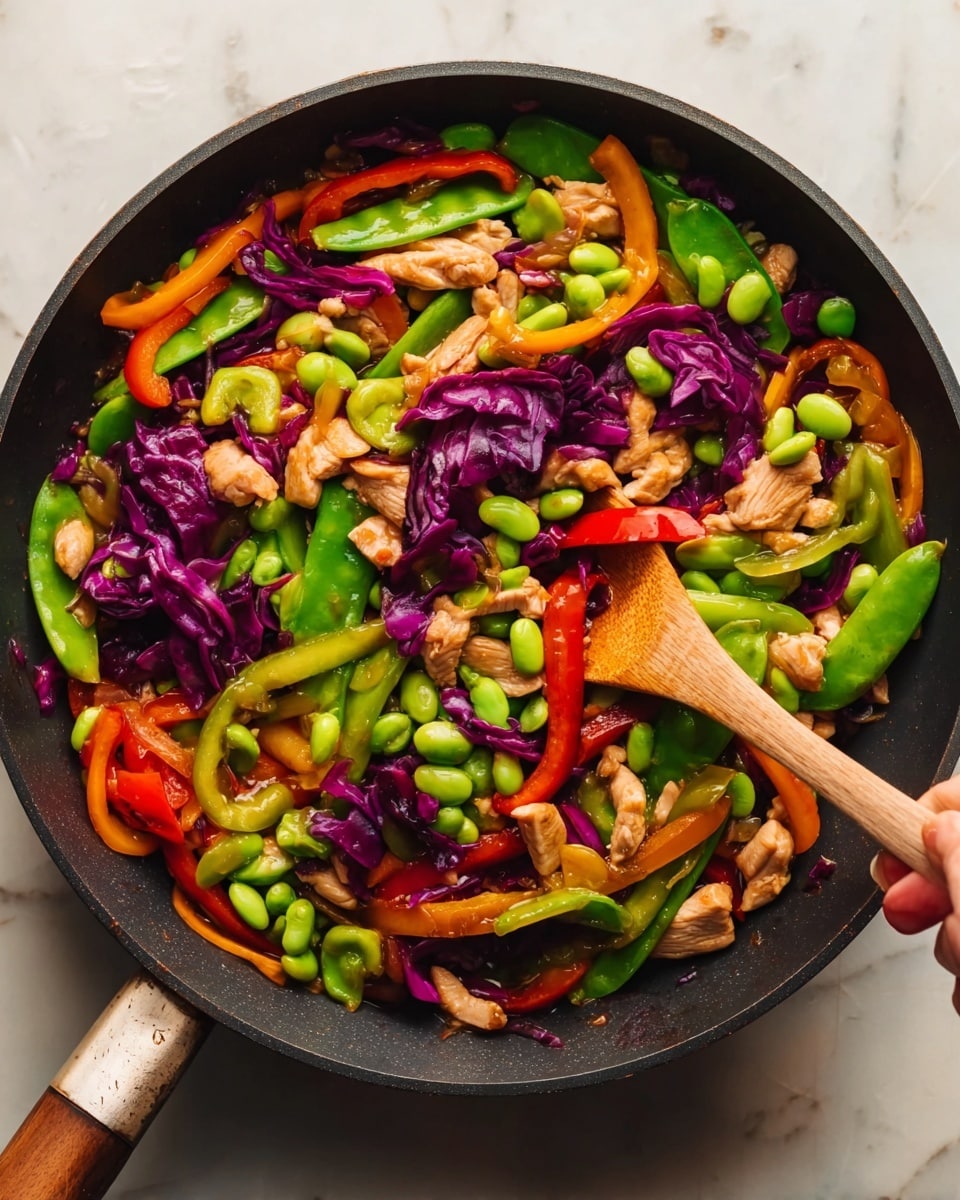 A black pan filled with a colorful stir-fry dish showing several layers of vibrant vegetables and pieces of chicken. The base consists of mixed bright green snap peas and edamame, creating a fresh and lively look. On top of that are thin slices of red, yellow, and orange bell peppers, adding rich, warm colors with a smooth texture. There are also chunks of purple cabbage giving a rough, leafy appearance, scattered throughout. The chicken pieces are golden-brown with a slightly crispy texture, arranged evenly across the pan. A woman's hand is holding a wooden spatula stirring the dish, with the pan sitting on a white marbled surface. Photo taken with an iphone --ar 4:5 --v 7