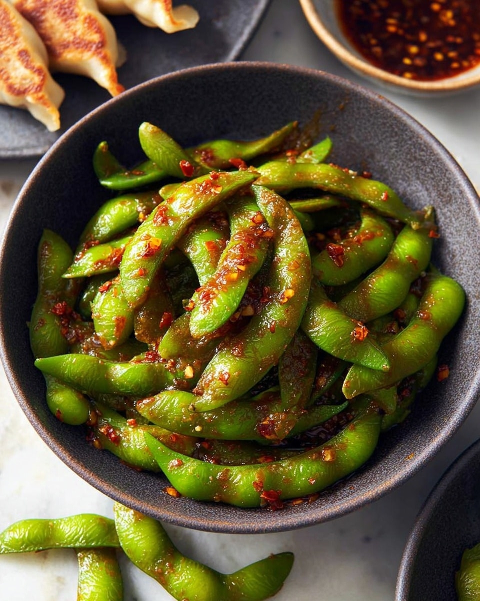 A close-up image shows a dark bowl filled with glossy green edamame pods coated in a reddish-brown spicy sauce scattered with chili flakes and seeds, the pods are piled high, some overlapping with smooth, slightly wrinkled textures visible; around the bowl, on a white marbled surface, a few edamame pods are scattered loosely, and another dark plate in the corner holds more of the sauced green pods, while in the blurred background, a white plate with several light golden-brown dumplings is visible, all items arranged in a cozy, casual setting. photo taken with an iphone --ar 4:5 --v 7