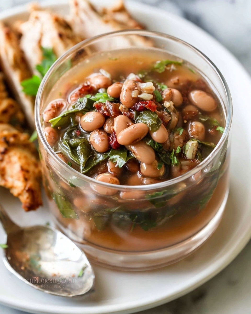 A clear glass bowl filled with cooked brown beans mixed with small pieces of green herbs and diced tomatoes, with a thick broth visible among the beans; the beans pile up about three layers deep, with a glossy and soft texture. The bowl sits on a white plate, next to pieces of cooked light brown chicken and a silver spoon on the right side. The white marbled surface beneath adds a clean, bright background. Photo taken with an iphone --ar 4:5 --v 7