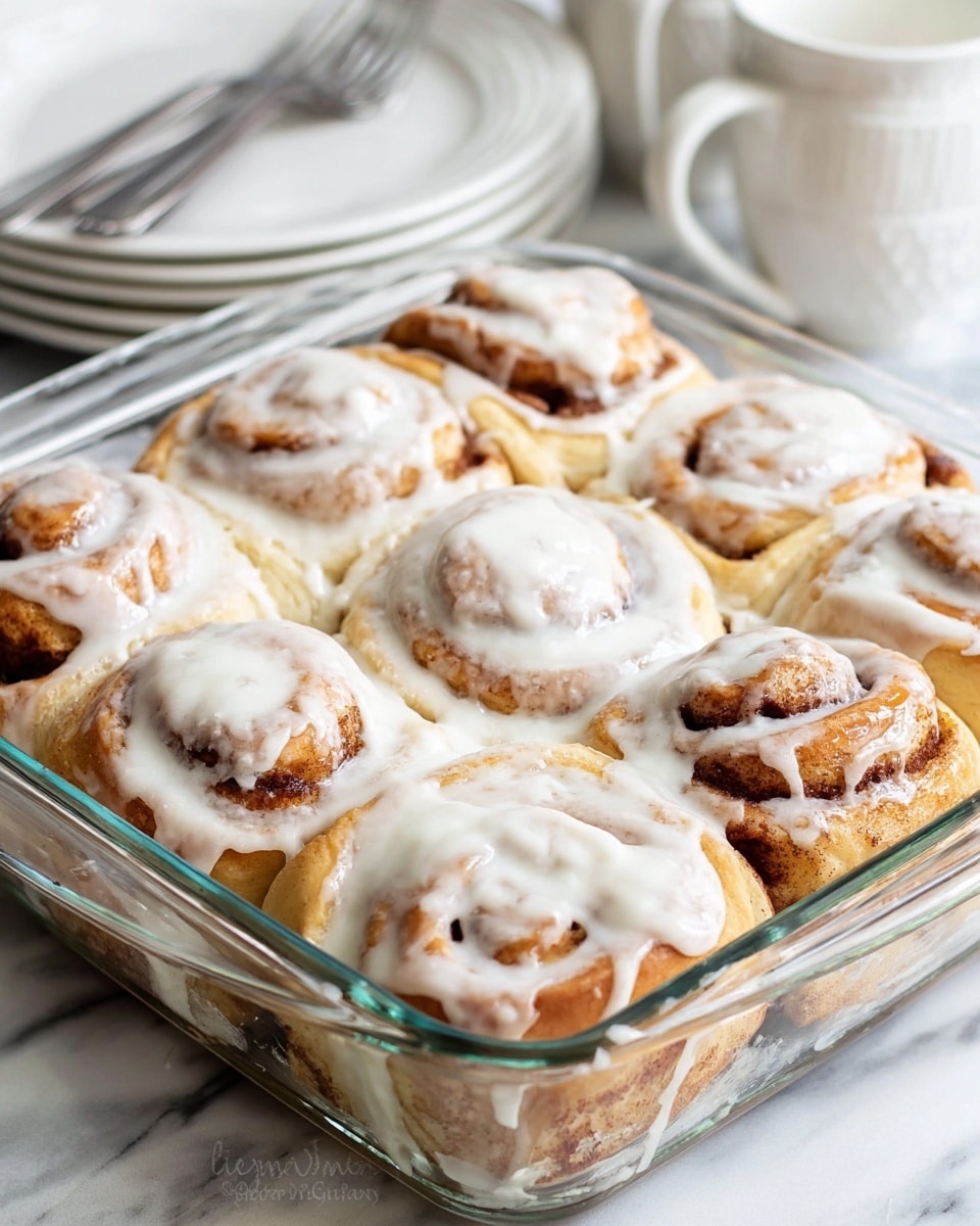 The image shows a glass baking dish filled with a dozen soft cinnamon rolls. Each roll has a golden-brown dough base with a dark cinnamon swirl visible in the spirals. They are covered with a thick, creamy white icing that glistens and drips slightly over the edges and between the rolls. The rolls are close together, filled tightly in the rectangular dish, creating a layered look with the dough's smooth texture underneath the thick glaze. The dish is set on a white marbled surface with some stacked white plates and a blurred background. photo taken with an iphone --ar 4:5 --v 7