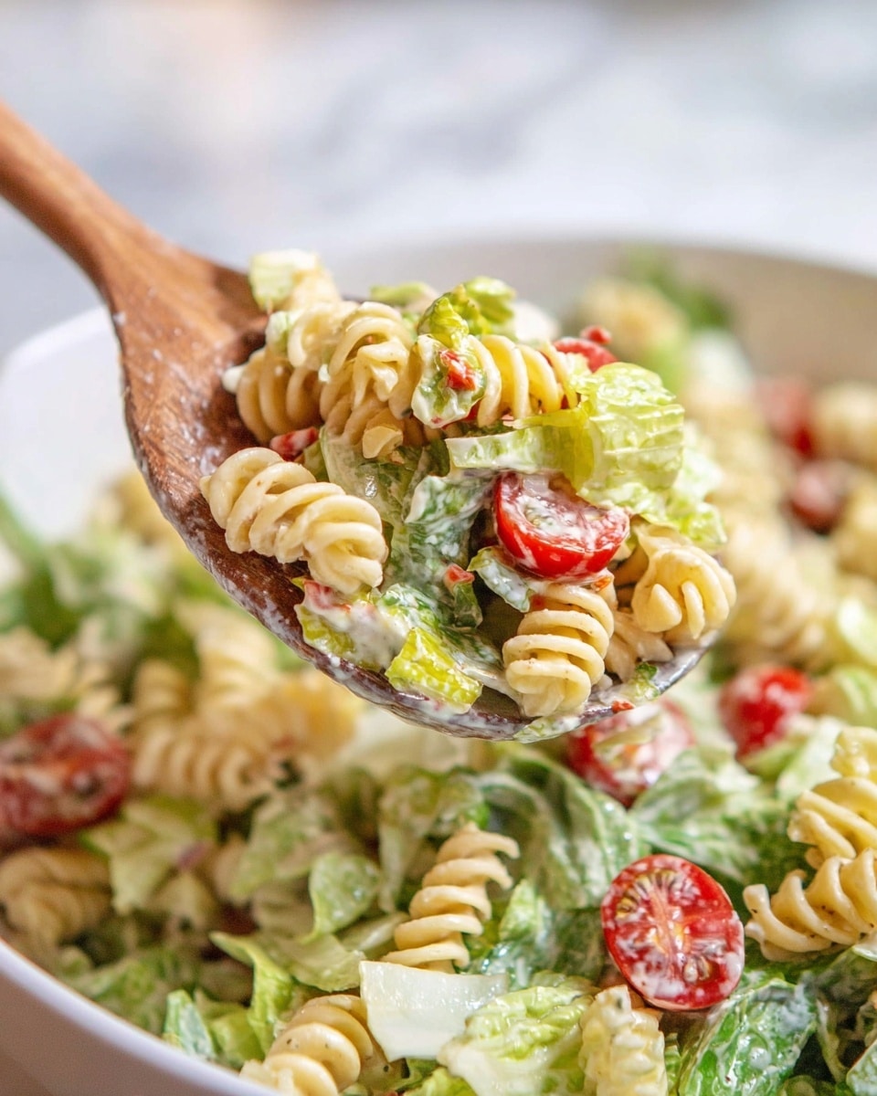 A close-up of a white bowl filled with rotini pasta salad, showing layers of twisted, light beige pasta spirals mixed evenly with bright green chopped lettuce leaves and small red cherry tomato halves. There are also bits of bacon or similar small brown pieces and creamy dressing coating the salad, giving a shiny texture. A wooden spoon lifts a portion of the salad, focusing on the mix of pasta, lettuce, and tomatoes. The background and surface consist of a white marbled texture. photo taken with an iphone --ar 4:5 --v 7