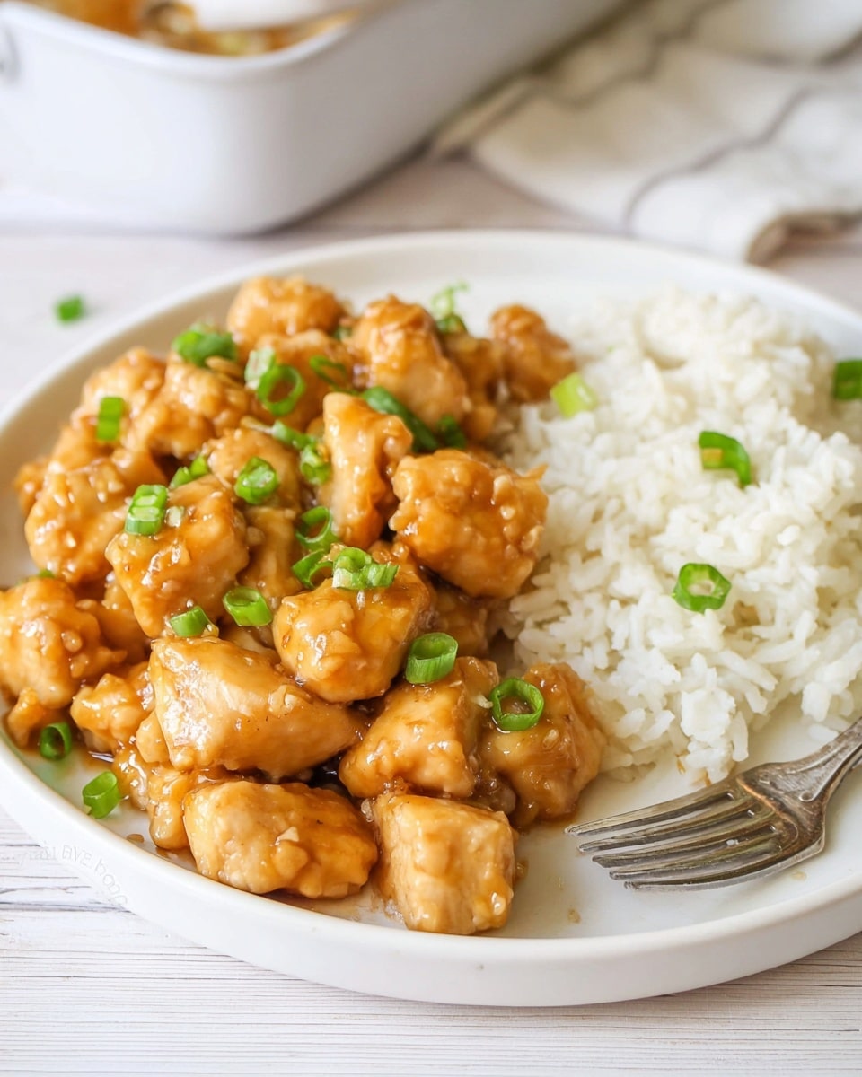 A white plate filled with two main layers, the left side showing a pile of golden-brown chicken pieces coated with a shiny, sticky light brown sauce and sprinkled with small green chopped scallions for color contrast, while the right side shows a mound of fluffy white rice mixed with a few scattered green scallion pieces, next to a silver fork resting on the plate’s edge, all placed on a wooden surface with a blurred white marbled texture background. photo taken with an iphone --ar 4:5 --v 7