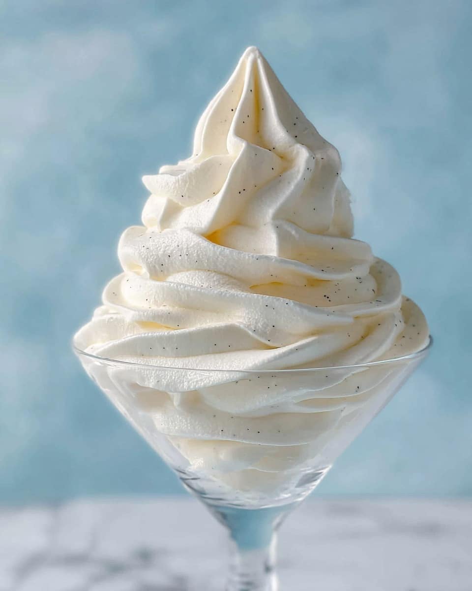 A tall swirl of whipped cream with soft, smooth texture is elegantly piped into a clear glass dessert cup. The whipped cream is bright white with tiny dark vanilla bean specks scattered throughout, creating subtle contrast. The layers of cream form a spiral that rises upward, with visible ridges and gentle curves that catch the light. The clear glass cup shows the cream’s texture and volume from the sides, while the background is a soft blue color with a white marbled texture beneath it. photo taken with an iphone --ar 4:5 --v 7