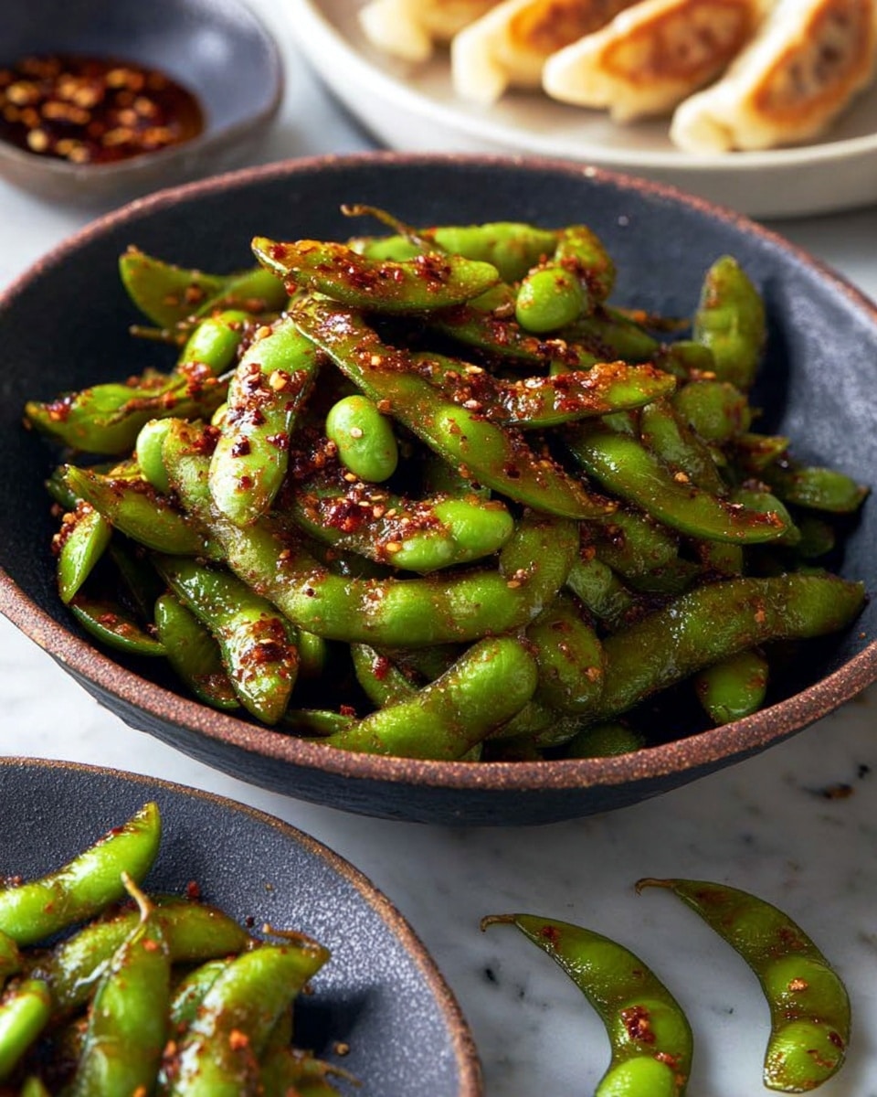 A close-up image shows a bowl filled with bright green edamame pods covered in a shiny, reddish-brown spicy sauce with small bits of chili and garlic. The bowl is dark gray and sits on a white marbled background. Around the bowl, a few edamame pods, also coated with sauce, are scattered on the surface. In the top left corner, part of a gray plate with browned dumplings is barely visible. The image has natural light that highlights the glossy texture of the sauce and the fresh look of the edamame. Photo taken with an iphone --ar 4:5 --v 7