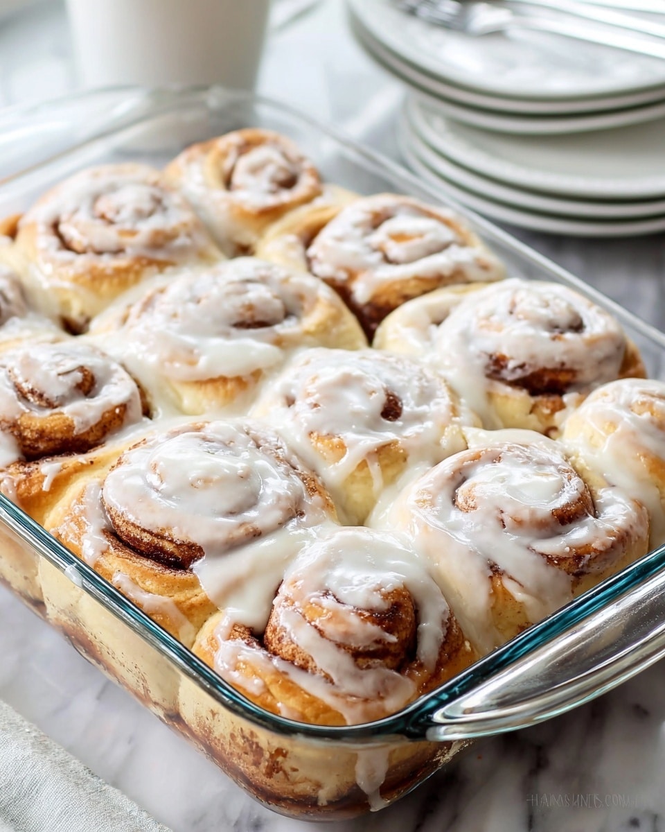 A clear glass rectangular dish holds nine soft, golden-brown cinnamon rolls arranged in three rows of three. Each roll shows spiraled layers of light brown dough mixed with darker cinnamon swirls. Thick, white icing covers the top of the rolls unevenly, with some drips running down the sides, adding a creamy texture. The background is a white marbled surface, and in the blurred background, a stack of white plates and a silver fork are barely visible. photo taken with an iphone --ar 4:5 --v 7