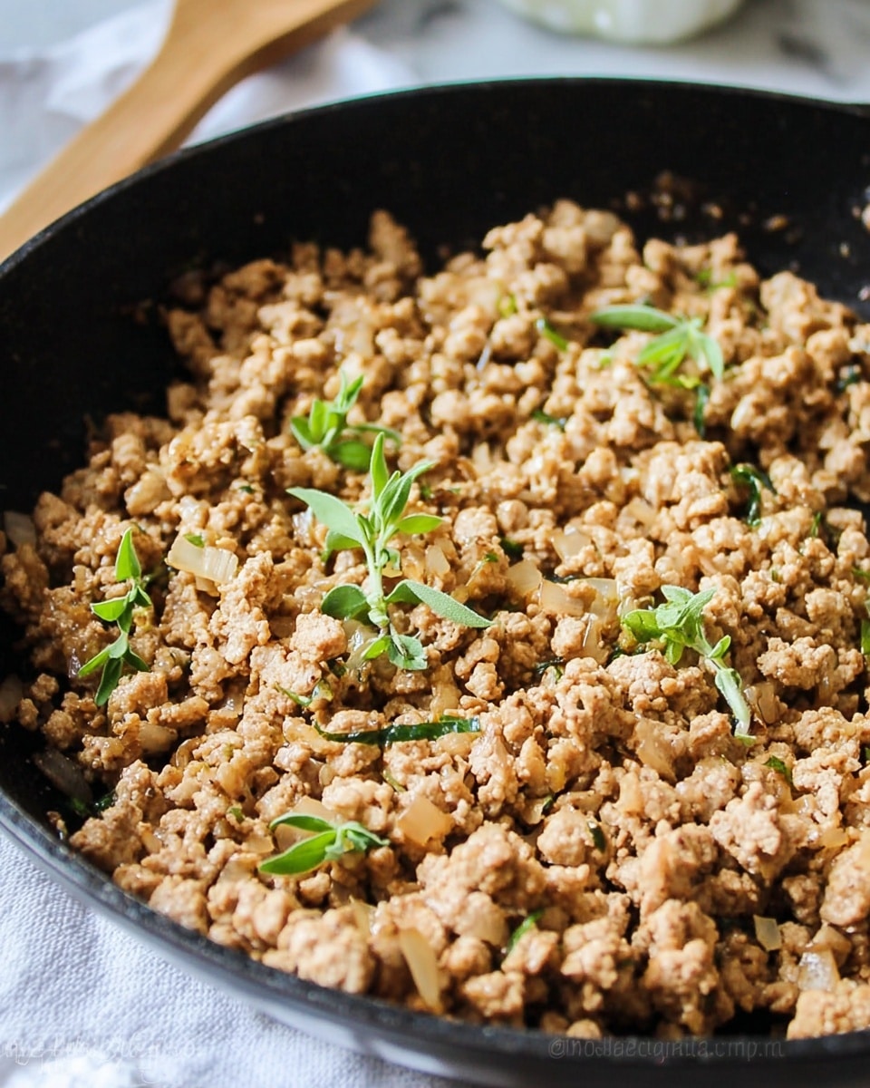 A close-up view of a black frying pan filled with cooked ground meat that is light brown and crumbly in texture. Small bits of translucent cooked onions are mixed in, and fresh green herb sprigs are scattered on top as garnish. The pan is set on a white marbled surface, with a wooden spoon partially visible to the side. The focus is clear on the meat and herbs, showing texture and small details. photo taken with an iphone --ar 4:5 --v 7