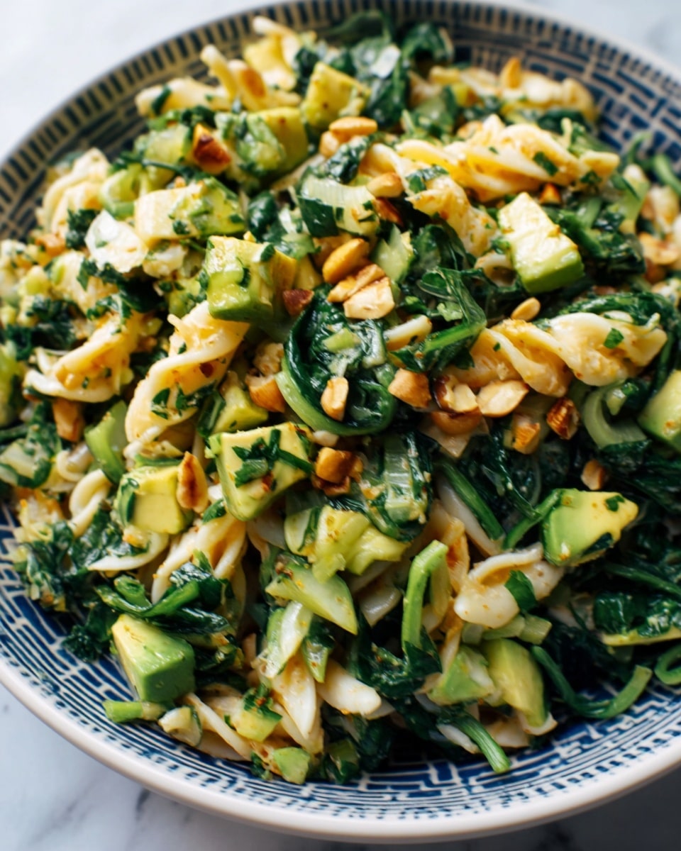 A close-up view of a pasta salad served in a white bowl with a gray pattern. The dish has twisted pasta pieces mixed with chopped green leafy vegetables and small chunks of light green avocado. Orange toasted nuts or seeds are scattered on top, adding texture and color contrast. The salad looks fresh and lightly dressed, with a mix of soft and crunchy elements. The bowl is placed on a white marbled surface. photo taken with an iphone --ar 4:5 --v 7