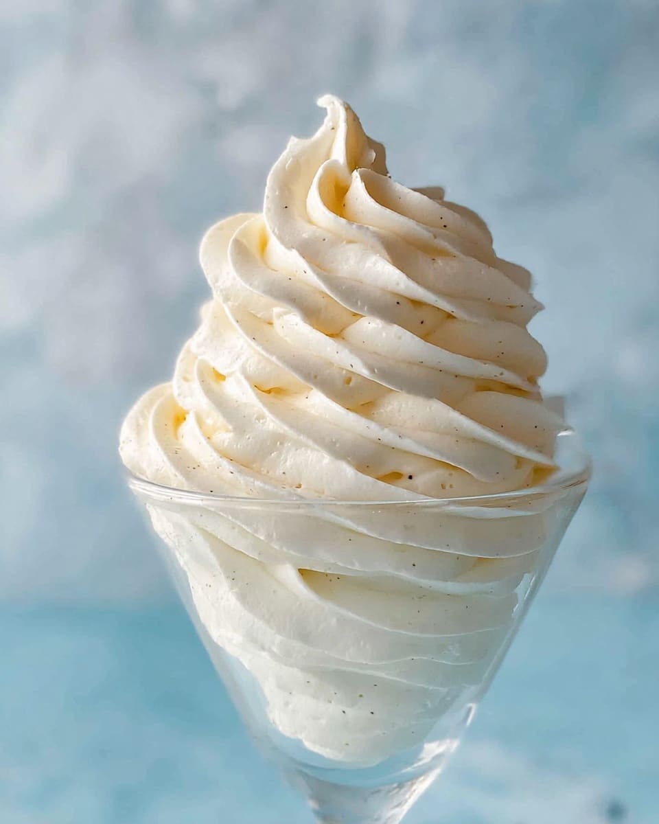 A clear glass dessert cup holds multiple swirls of soft, creamy white whipped cream, stacked in thick, smooth layers that rise to a tall, pointed peak with gentle curves and ridges, showing tiny black vanilla bean specks throughout. The smooth, shiny texture of the whipped cream contrasts with the transparent, simple glass, all set against a soft blue background with a subtle out-of-focus effect, placed on a white marbled surface. Photo taken with an iphone --ar 4:5 --v 7
