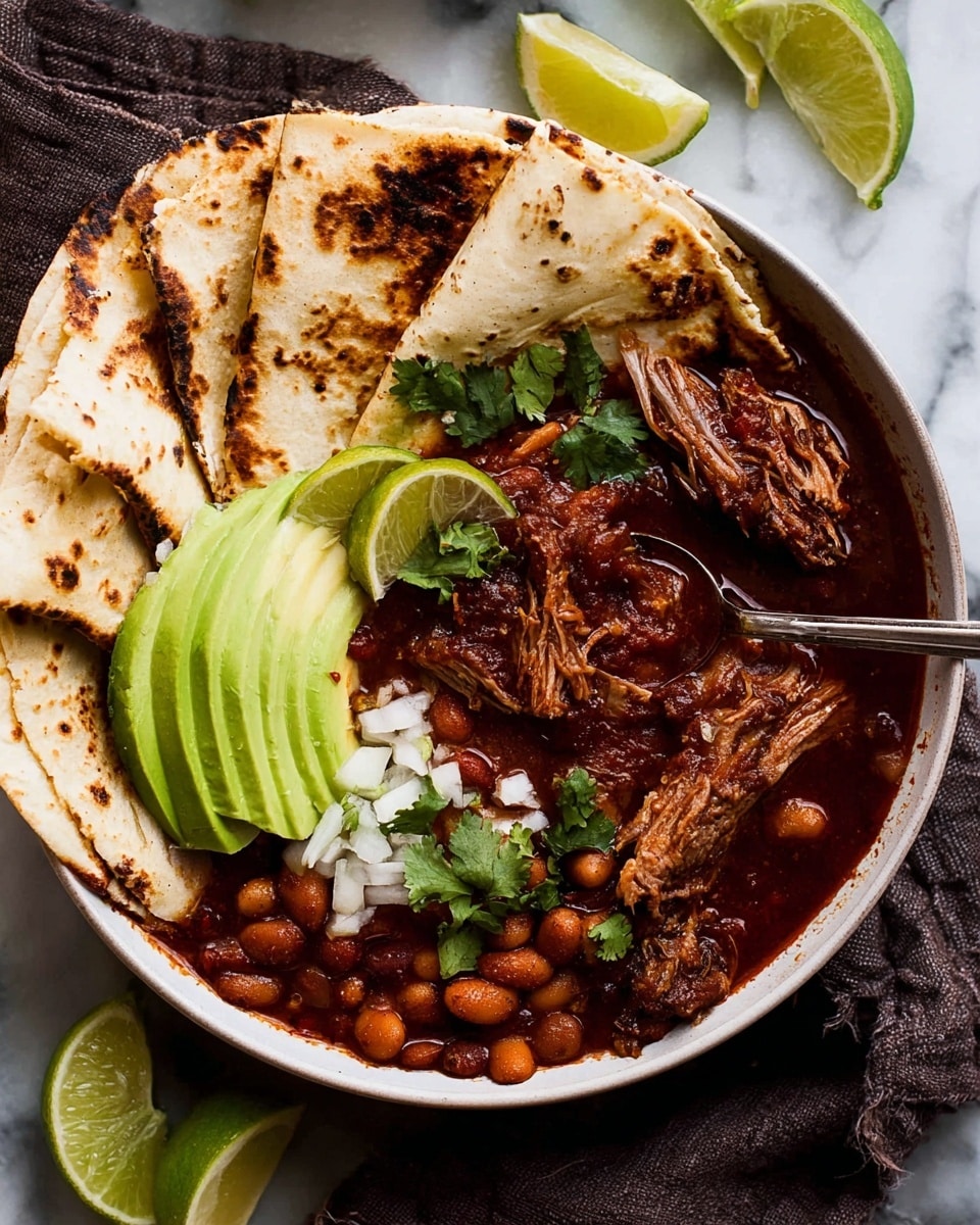 A white bowl holds a rich, dark red stew with chunks of shredded meat and soft beans, covered in thick sauce, topped with green cilantro leaves. On one side inside the bowl, there are thin slices of bright green avocado sprinkled with small white onion pieces. Next to the avocado, several folded pieces of lightly charred tortilla rest along the edge of the bowl. Two lime wedges, one green and one yellow, are placed on top of the tortillas. A silver fork is pulling apart a piece of meat in the stew. Around the bowl, more folded charred tortillas and lime wedges lie on a white marbled surface. Photo taken with an iphone --ar 4:5 --v 7