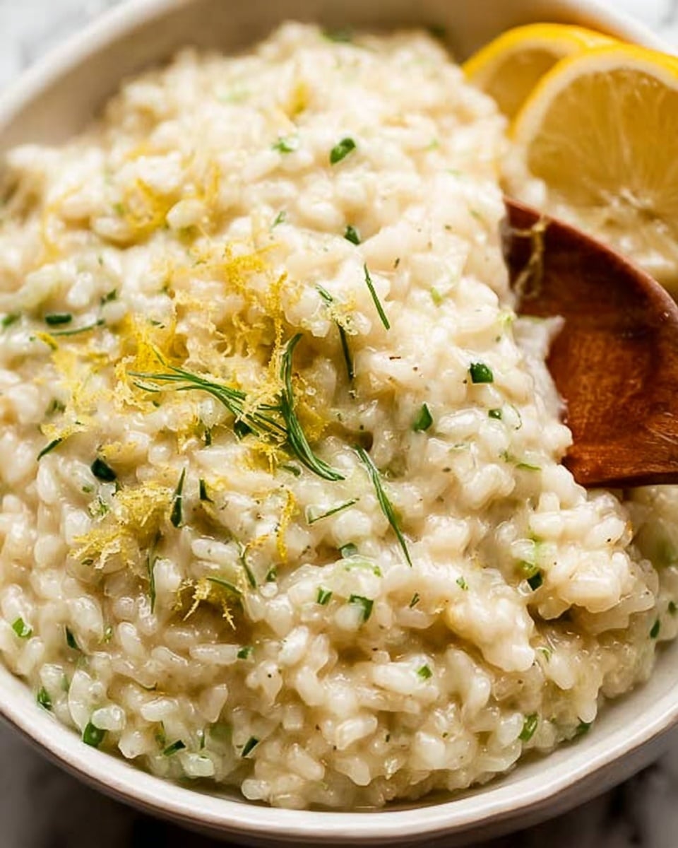 A close-up view of creamy risotto in a white bowl filled almost to the top, with soft, individual grains of rice visible in a thick, smooth sauce. The risotto is pale yellow with small bits of fresh green herbs sprinkled evenly throughout, adding a touch of color. Small pieces of lemon zest are scattered on the surface, and a few thin lemon slices rest slightly buried in the risotto. A wooden spoon is partially submerged on the right side, mixing the dish. The background is a white marbled texture. photo taken with an iphone --ar 4:5 --v 7