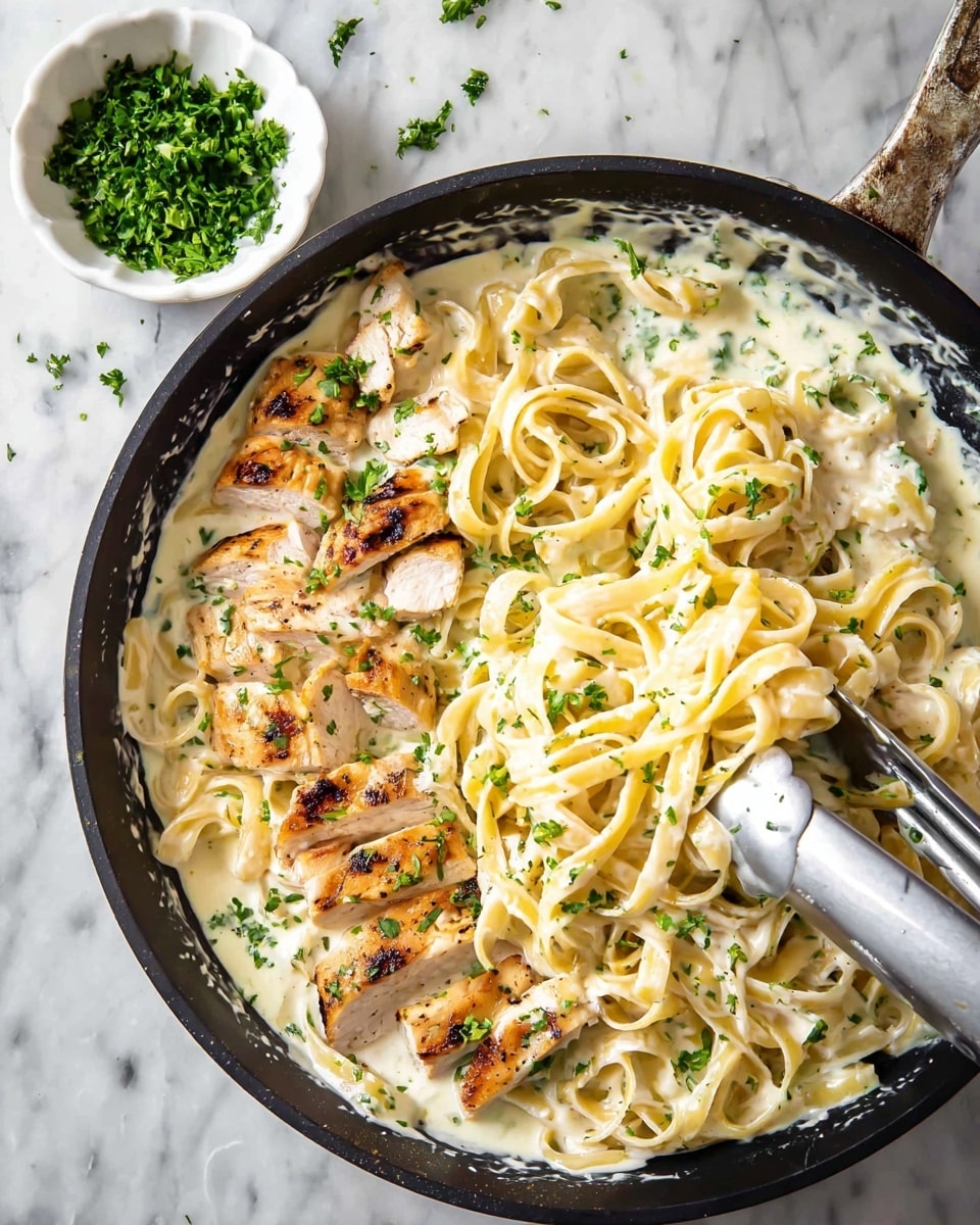 In a black pan on a white marbled surface, there is a creamy white sauce coating the whole dish. On top of the sauce, light golden fettuccine pasta is mixed in thick curls. Thin slices of cooked chicken with light browned edges are scattered evenly throughout the pasta. Small pieces of fresh green herbs are sprinkled across the dish, adding color contrast. To the top left, a white bowl filled with finely chopped green herbs sits. A pair of silver tongs holds some pasta and chicken in the center of the pan. photo taken with an iphone --ar 4:5 --v 7